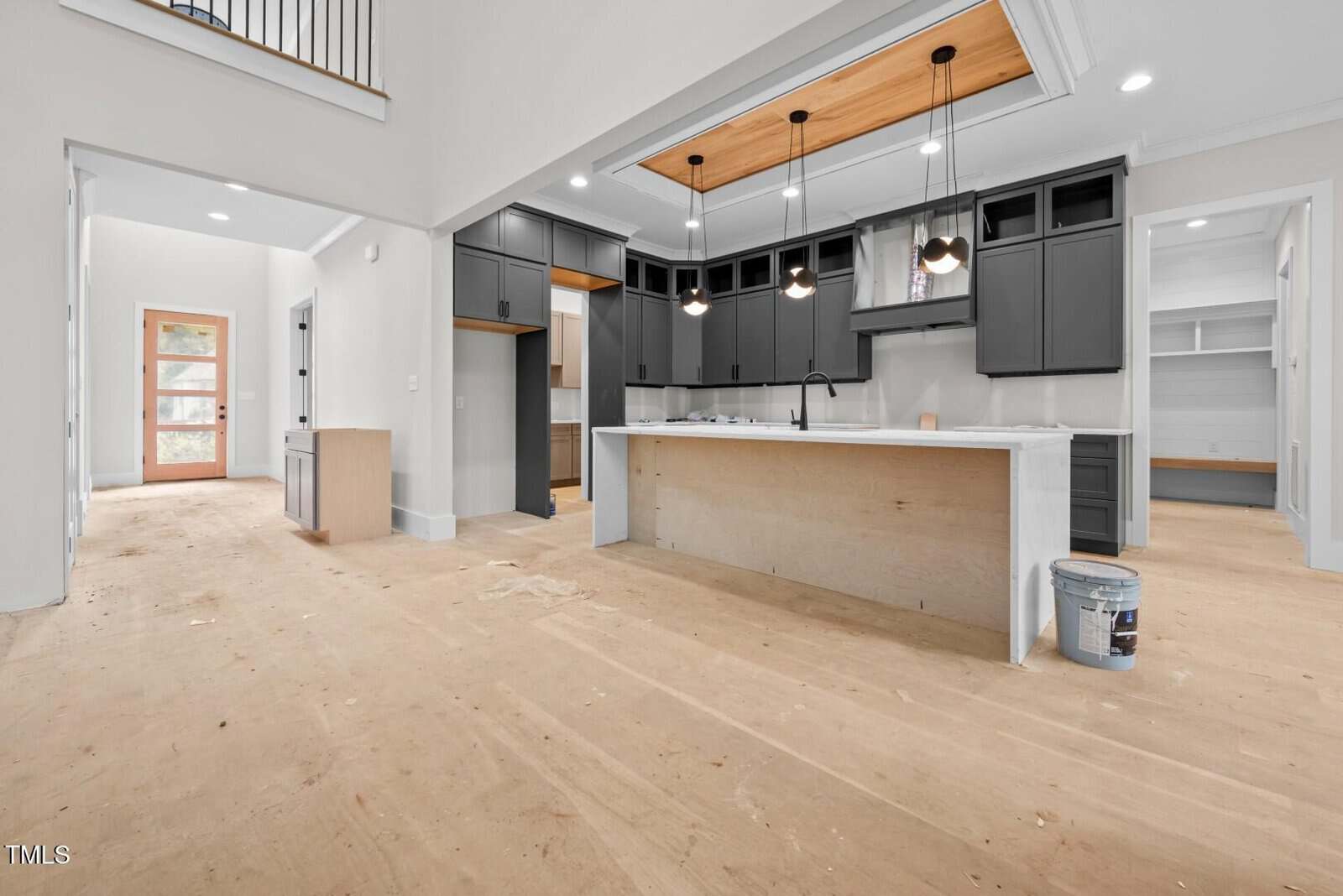 A modern, unfinished kitchen with black cabinets, white countertops, and pendant lights above the island. The floor is bare with construction materials visible, and an open doorway leads to other rooms.