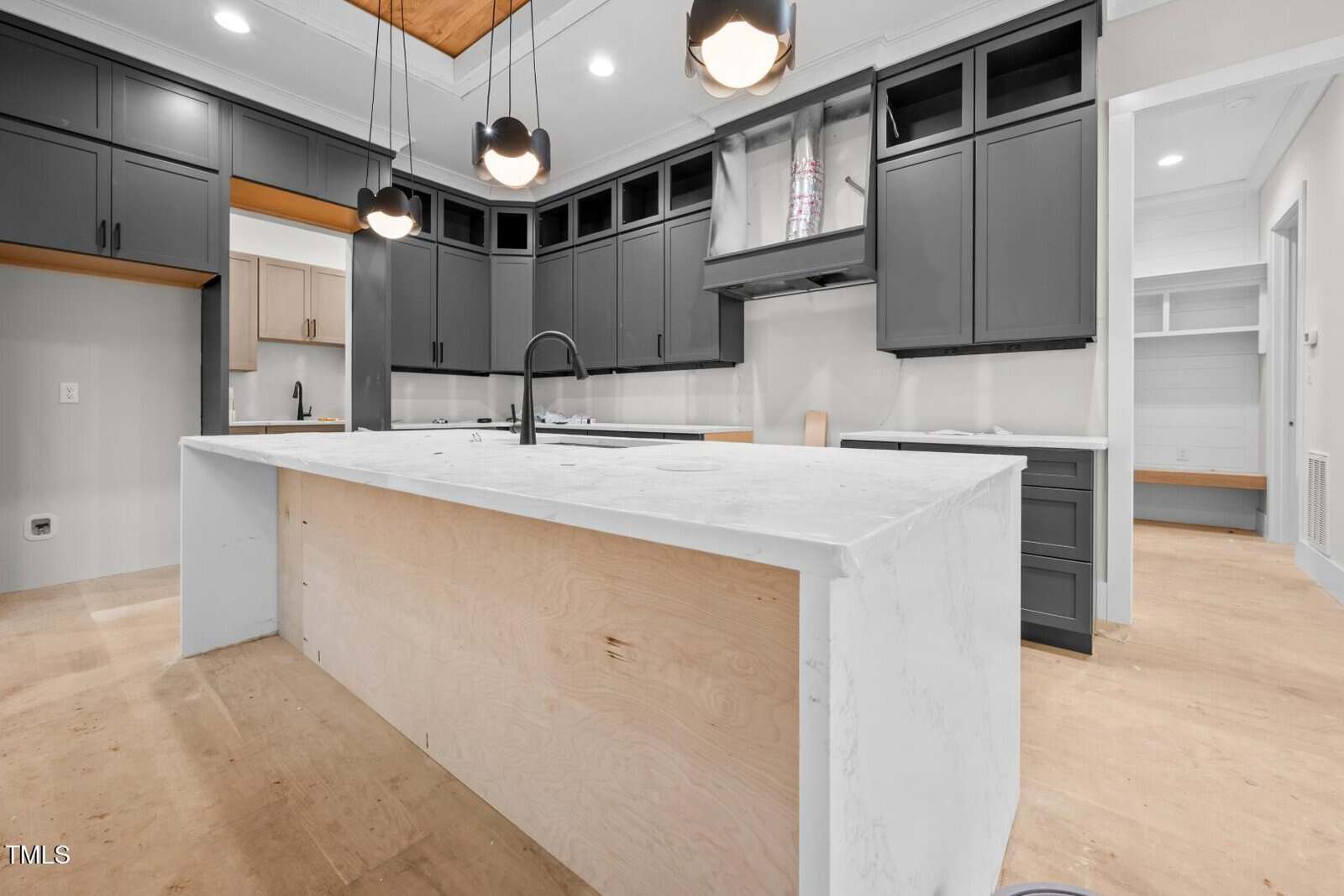A modern kitchen under construction featuring dark gray cabinets, a large white marble island without drawers or panels, pendant lights overhead, and unfinished floors. Some areas lack hardware and appliances.
