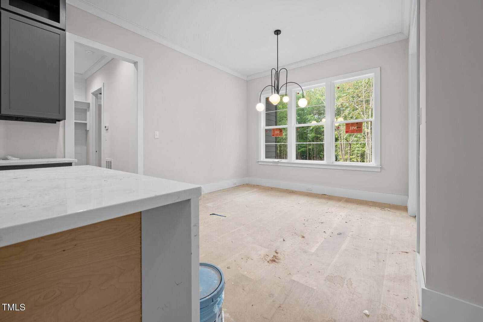 A partially finished room with bare floors, a blue paint bucket, and a white countertop in the foreground. A modern light fixture hangs from the ceiling, and two large windows overlook green trees outside.