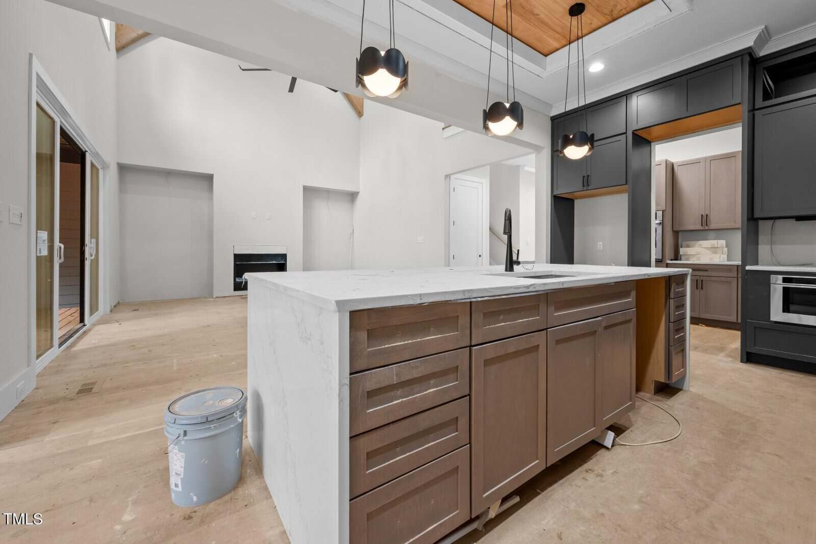 A modern kitchen under construction features a large island with a marble countertop, brown cabinets, hanging pendant lights, and unfinished floors. A paint bucket sits on the bare floor near the island.