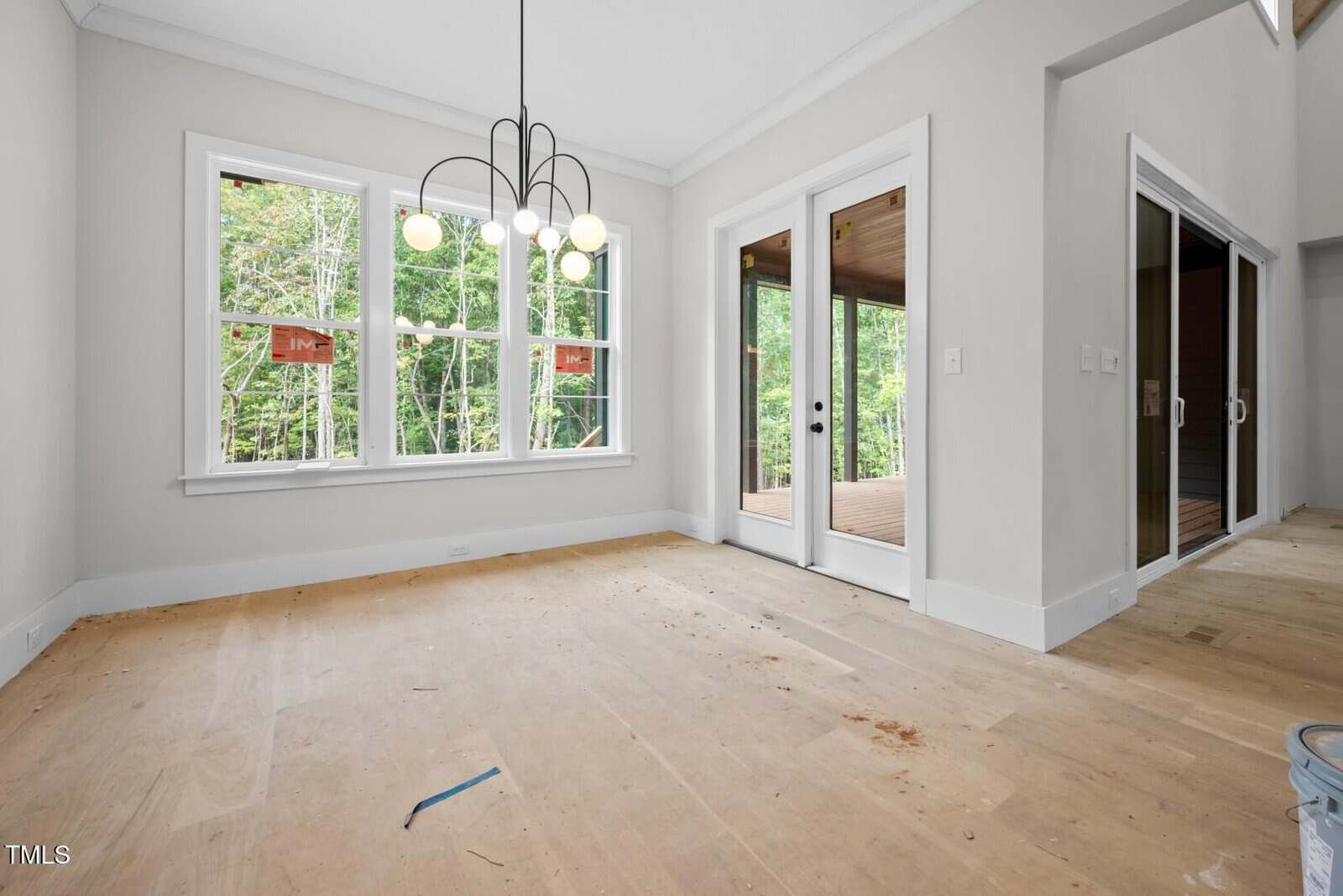 An unfurnished room with bare plywood floors, large windows with trees visible outside, a modern chandelier, and glass double doors leading to a covered porch. White trim and light-colored walls are present throughout.