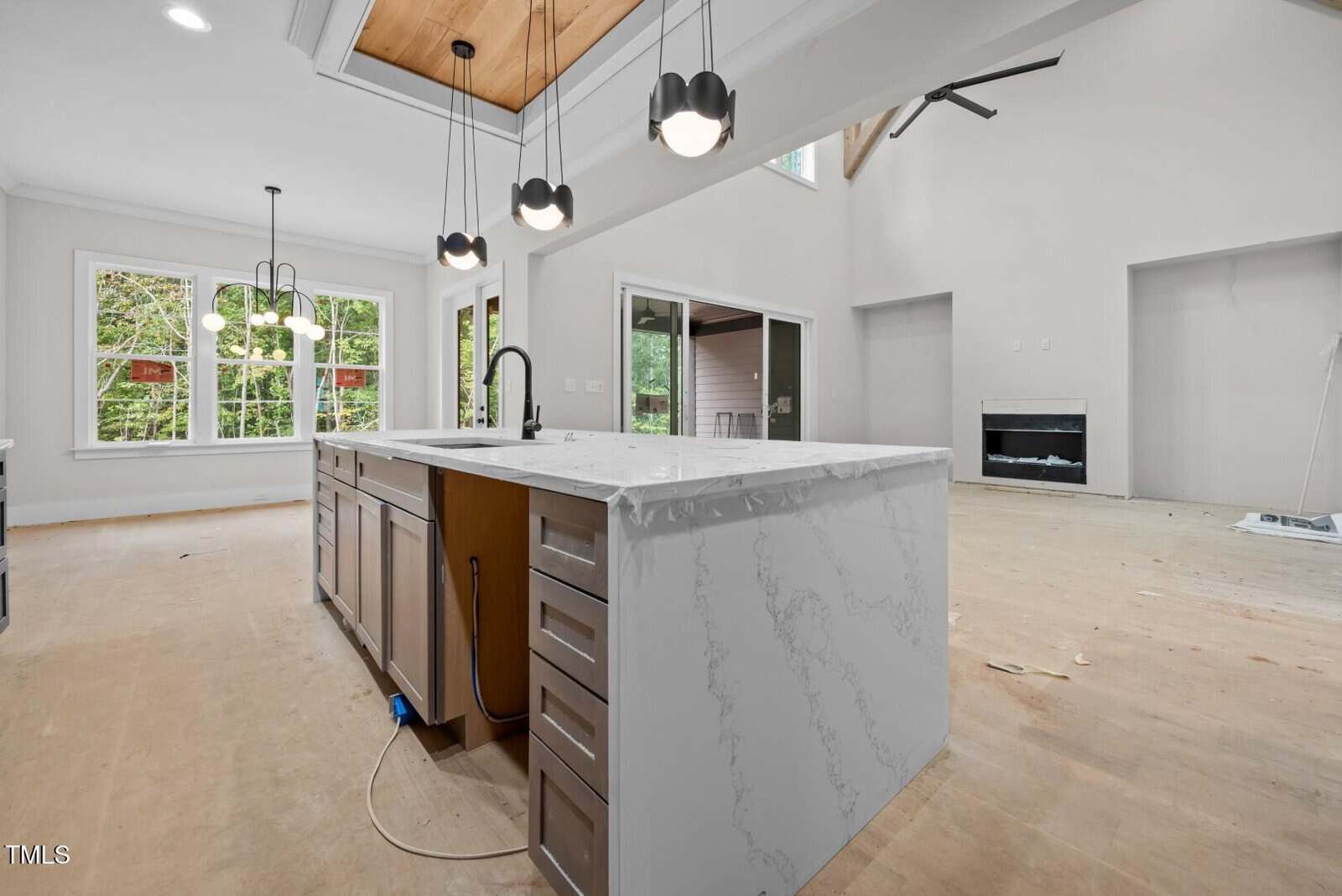 A modern, unfinished kitchen with a large marble island, pendant lights, and stainless steel sink. The floor is bare, and an open living area with a fireplace is visible in the background.