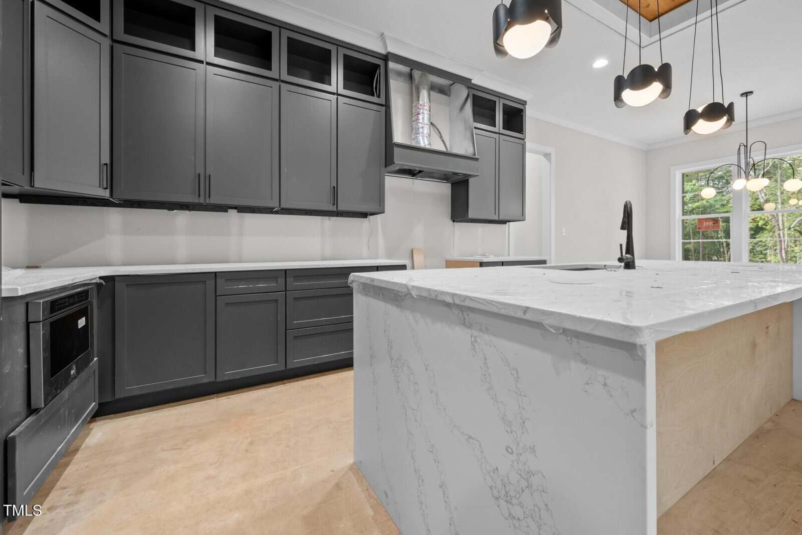 Modern kitchen with dark gray cabinets, white marble countertops, built-in oven, island with sink, pendant lights, and large window letting in natural light.