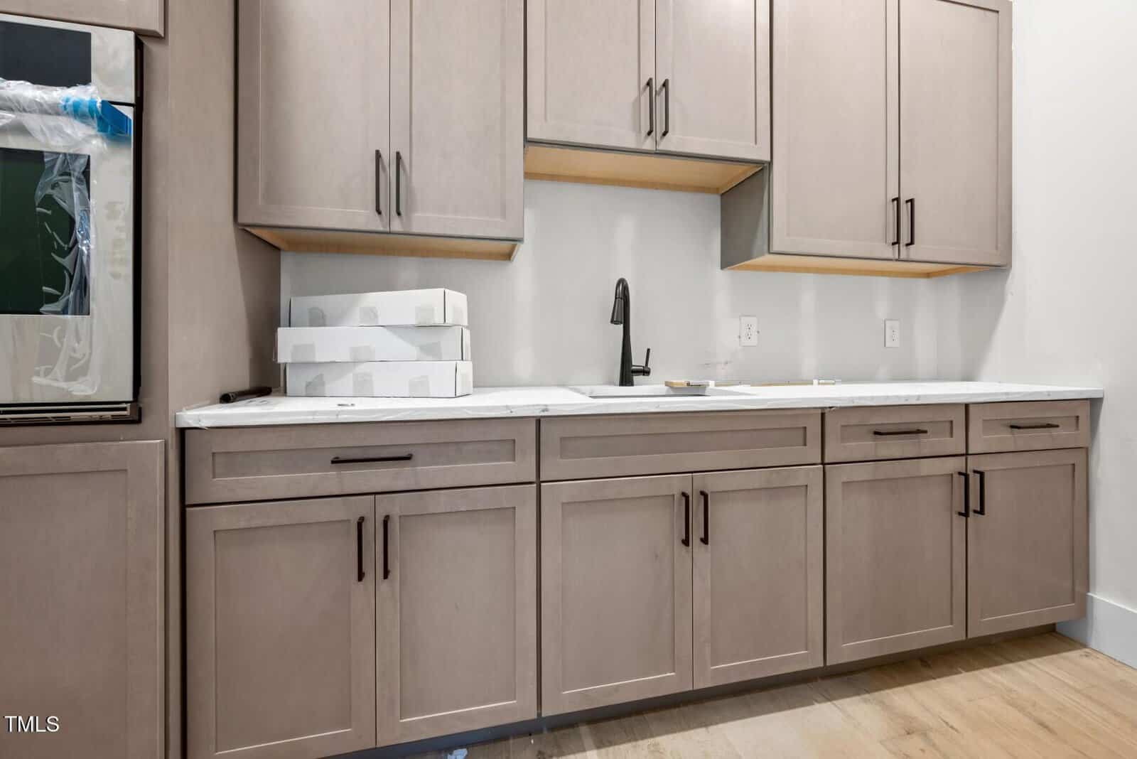 Modern kitchen with light wood cabinets, white marble countertop, matte black faucet, and three stacked white boxes on the counter under upper cabinets. Light wood flooring is visible below.