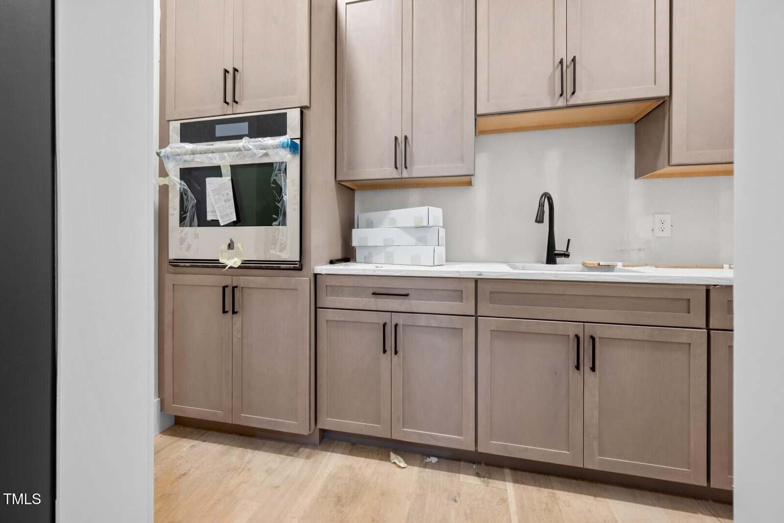 Modern kitchen with light wood cabinets, an oven covered in protective plastic, white countertops, a black faucet, and a set of white boxes on the counter. The room appears to be under construction or recently remodeled.