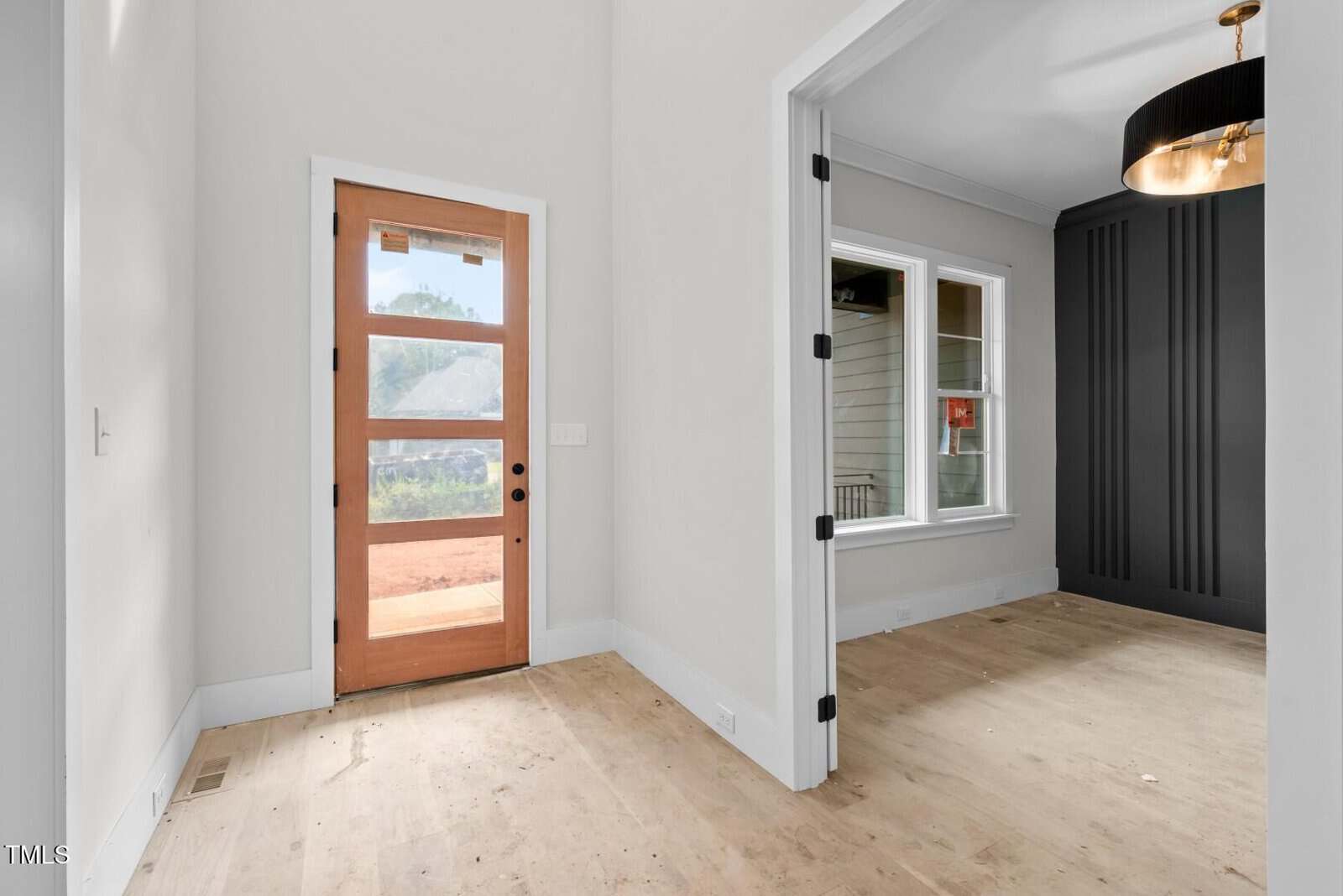 A view of a modern entryway with a wooden front door featuring glass panels, adjacent to a room with a large window, dark accent wall, and a ceiling light fixture, both with light-colored walls and floors.
