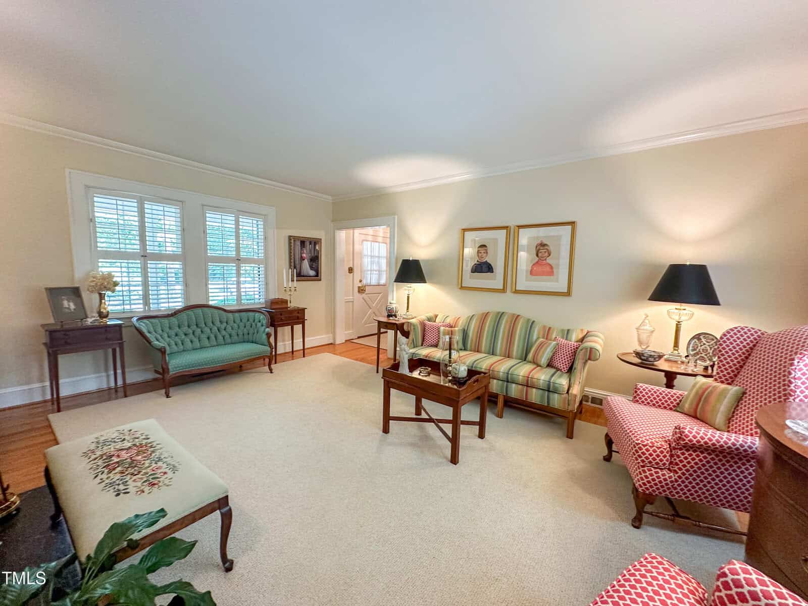 A cozy living room with vintage-style furniture, including striped and patterned armchairs, a green settee, wooden tables, lamps, framed portraits on the wall, and large windows with white shutters letting in natural light.