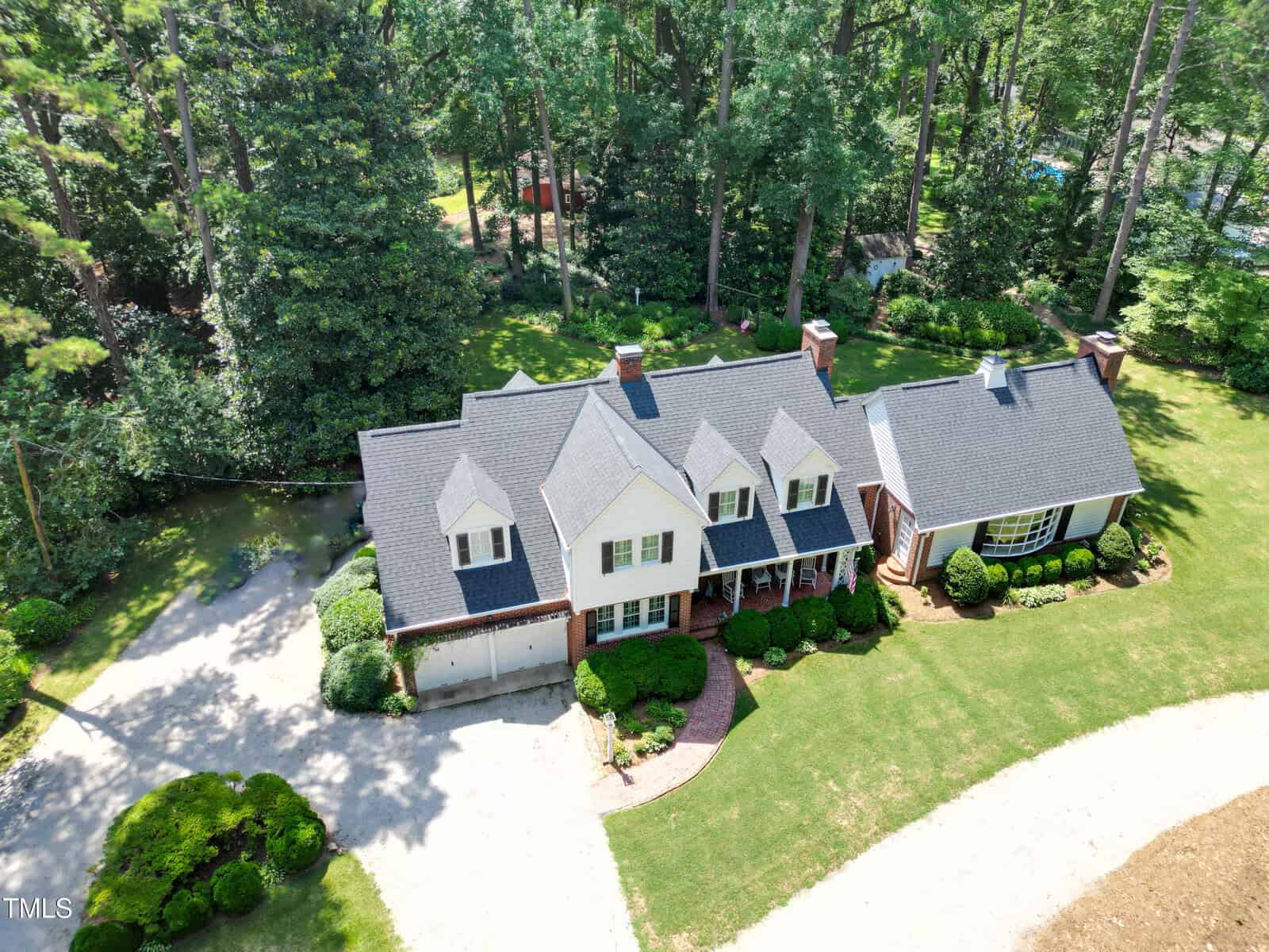 Aerial view of a large two-story house with gray roof, white and brick exterior, surrounded by trees, shrubs, and a curved driveway.