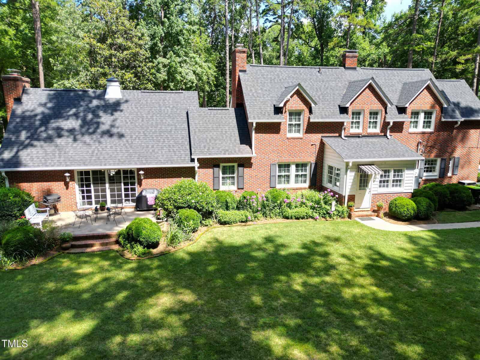 A large red brick house with dark shingles, multiple chimneys, white-framed windows, and a well-kept lawn surrounded by shrubs and trees on a sunny day.