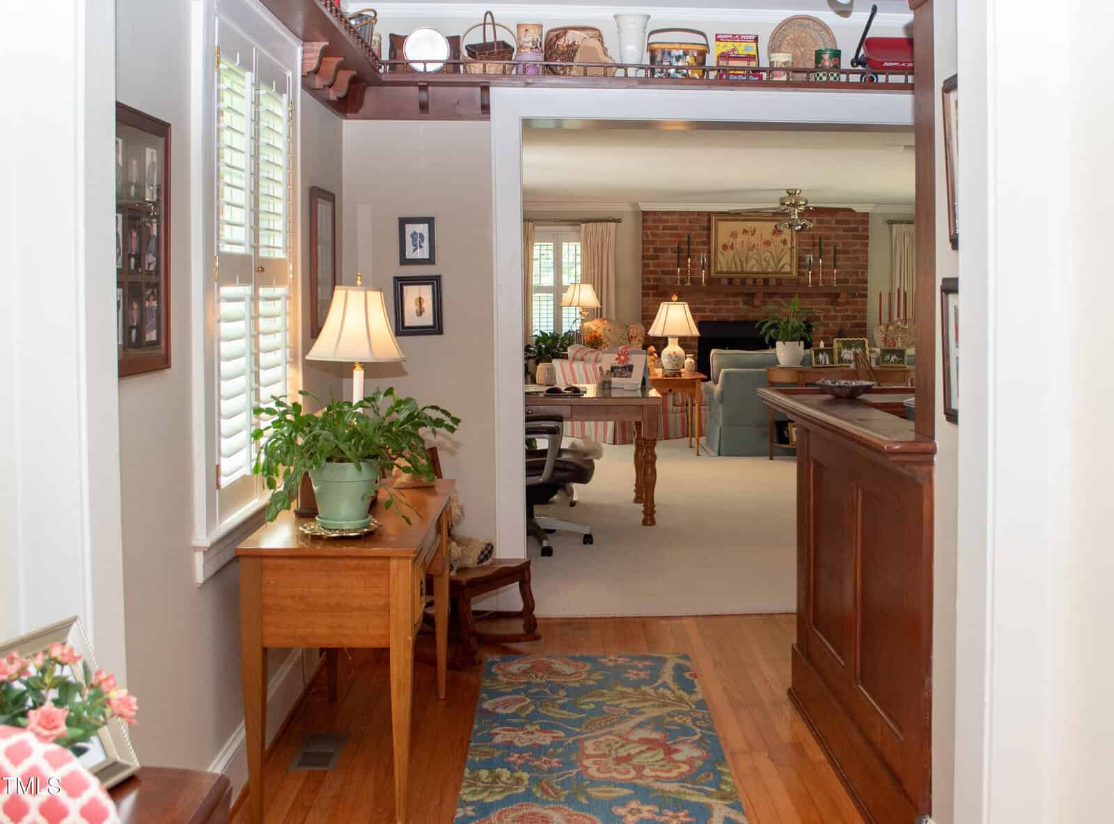 A hallway with a patterned rug, wooden floors, and a table with a lamp and plant leads to a cozy living room with a brick fireplace, armchairs, and decorative items on shelves and mantle.