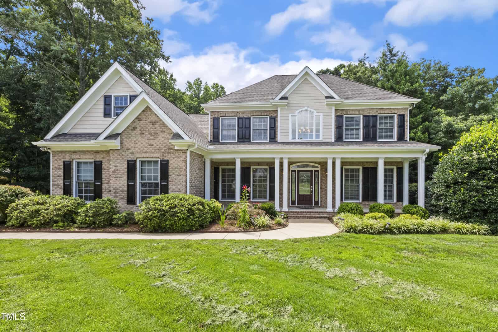 Two-story suburban house with brick and white siding exterior, black shutters, large front porch with white columns, manicured bushes, and a green lawn under a blue sky with clouds.