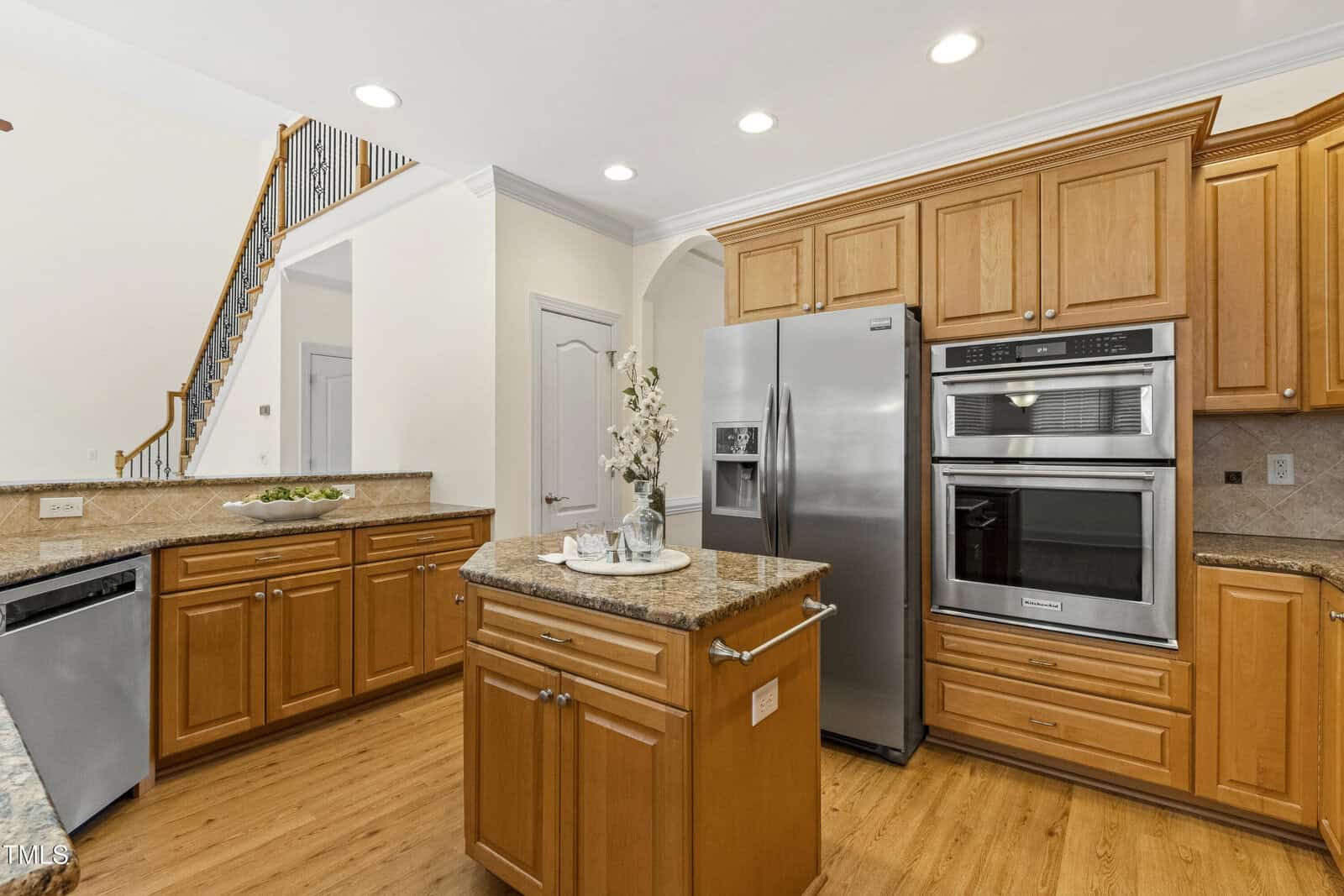 A modern kitchen with wood cabinets, stainless steel appliances, granite countertops, a small island with a floral arrangement, and stairs visible in the background. Recessed lighting brightens the space.