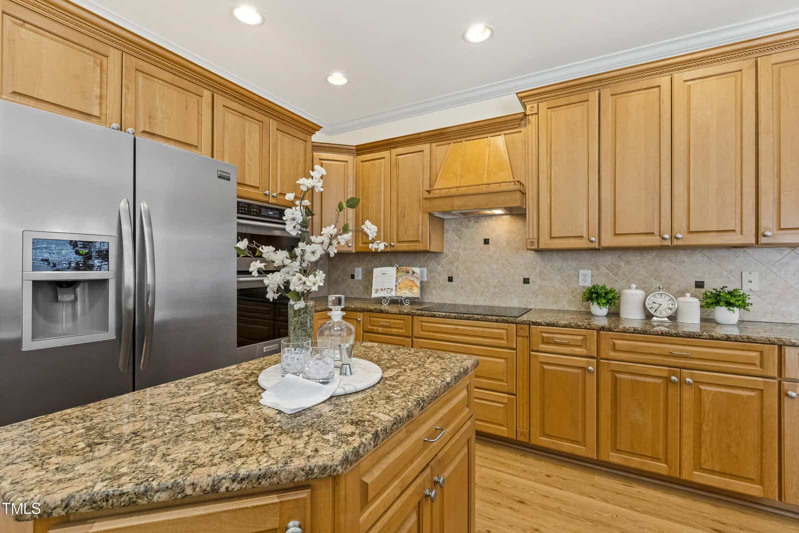 A spacious kitchen with light wood cabinets, stainless steel appliances, a granite island countertop with a flower vase, and decorative plants, all under bright recessed lighting.