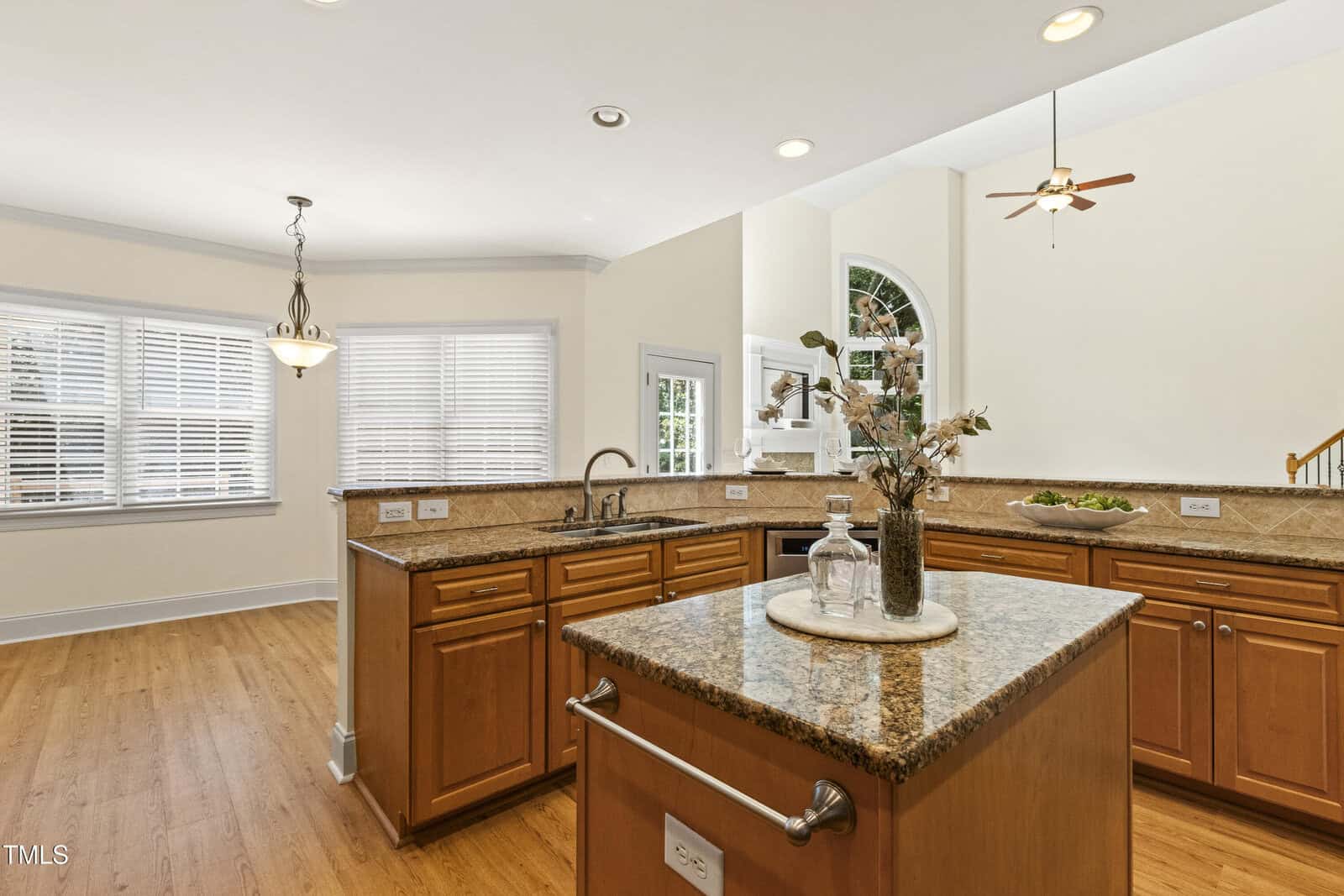 Spacious kitchen with wood cabinets, granite countertops, and a central island. Large windows provide natural light. Stainless steel appliances and a sink are visible. Light fixtures are mounted on the ceiling.