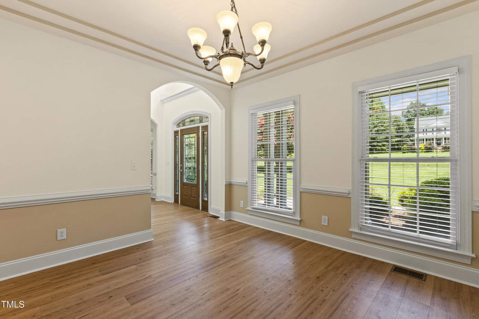 A dining room with wood floors, beige and white walls, white trim, large windows, and a glass-paneled double door entry. A chandelier hangs from the ceiling. Sunlight streams in through the windows.