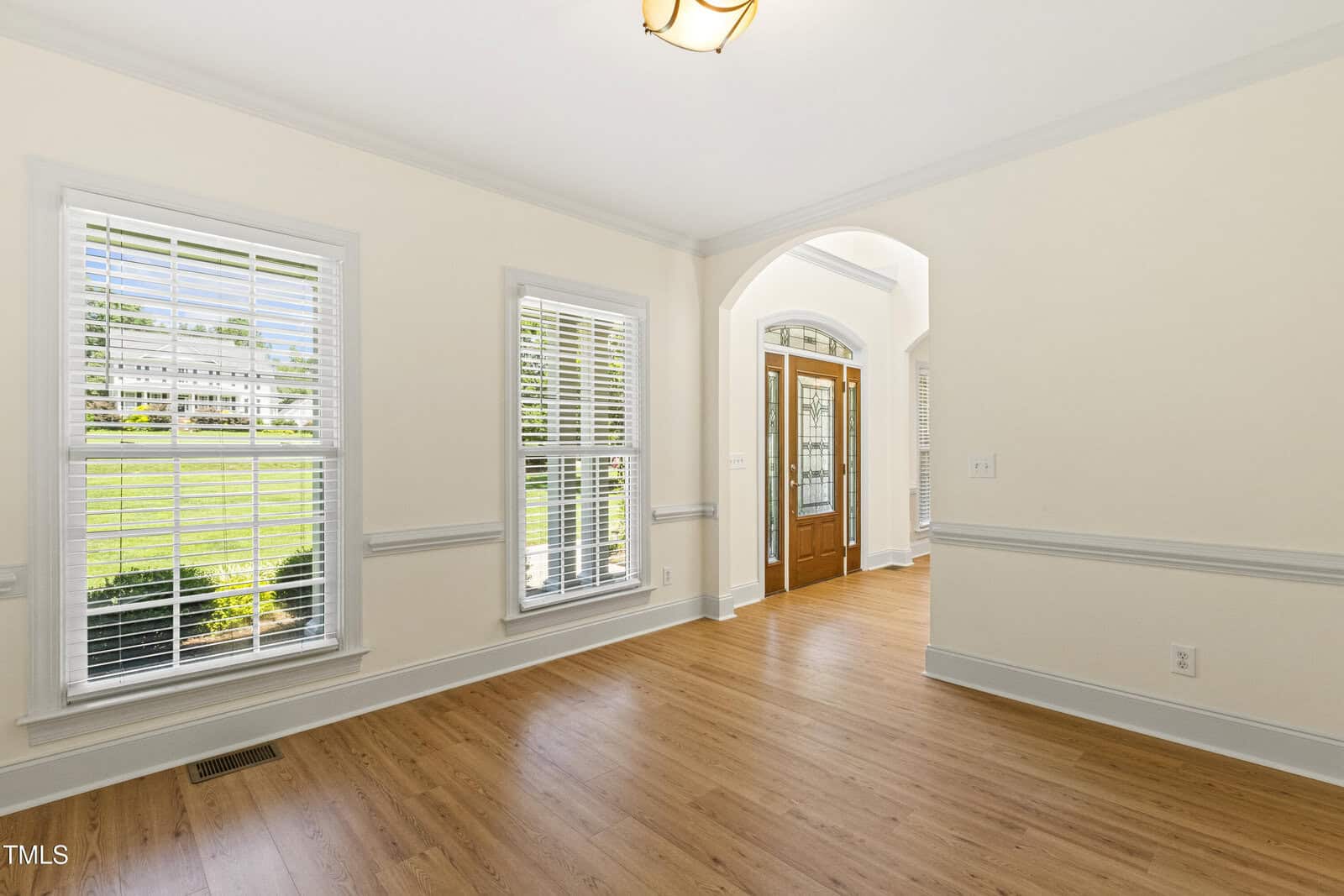 Bright room with light wood floors, cream walls, three large windows with white blinds, and a wooden front door with glass panels. Sunlight streams in, and green grass is visible outside. Trim and ceiling are white.