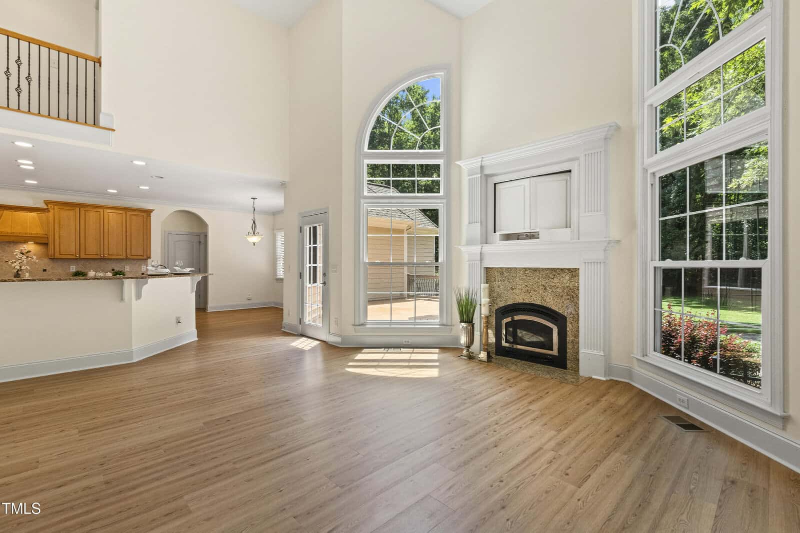 Spacious living room with tall ceilings, large arched windows, a white fireplace, light wood flooring, and an open view into a kitchen with wooden cabinets and a hanging light fixture. Lush greenery visible outside.