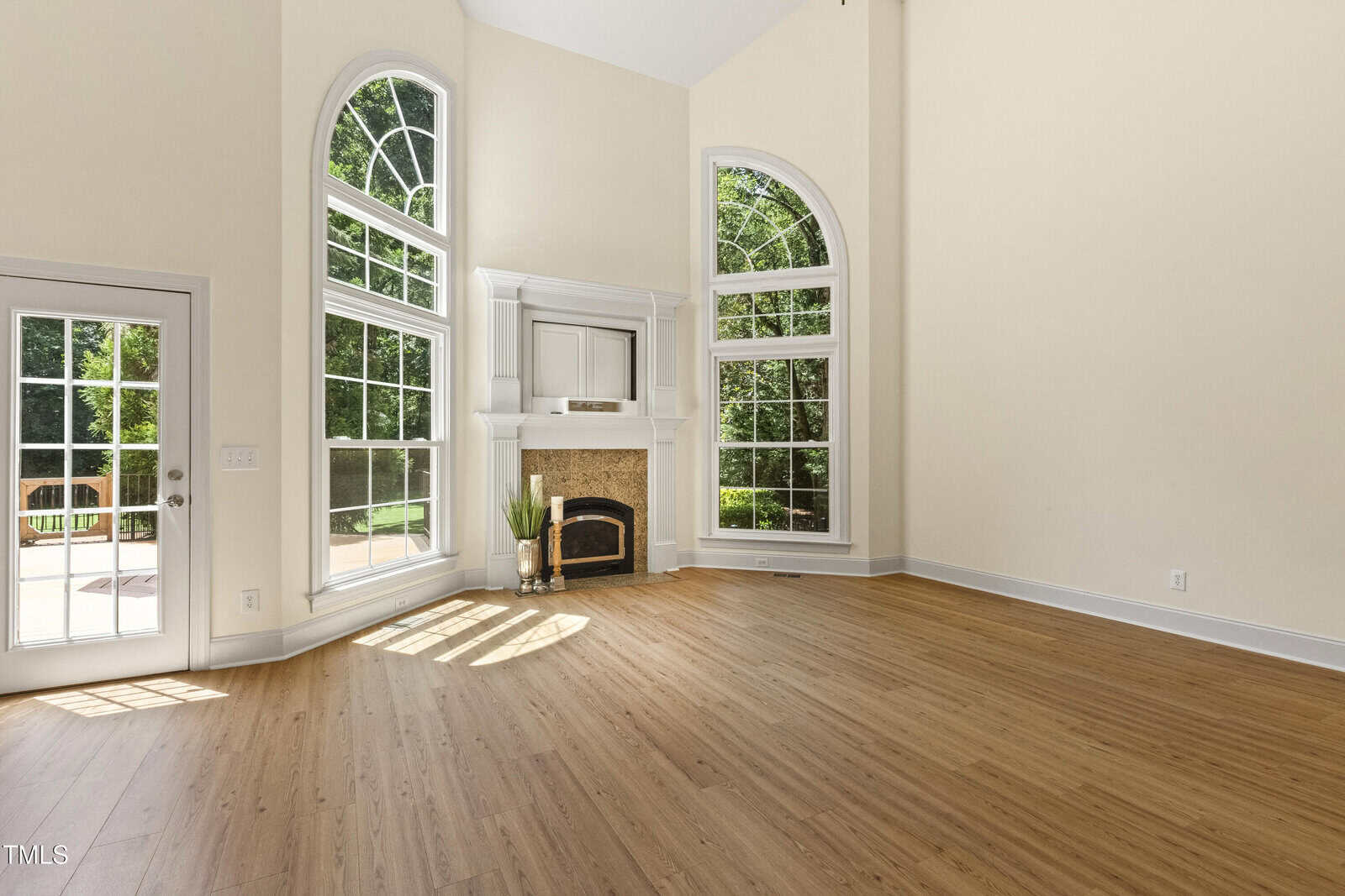 Bright living room with tall, arched windows, a glass door leading outside, and a fireplace with a white mantel. The space has light-colored walls and wood flooring, with sunlight streaming in.