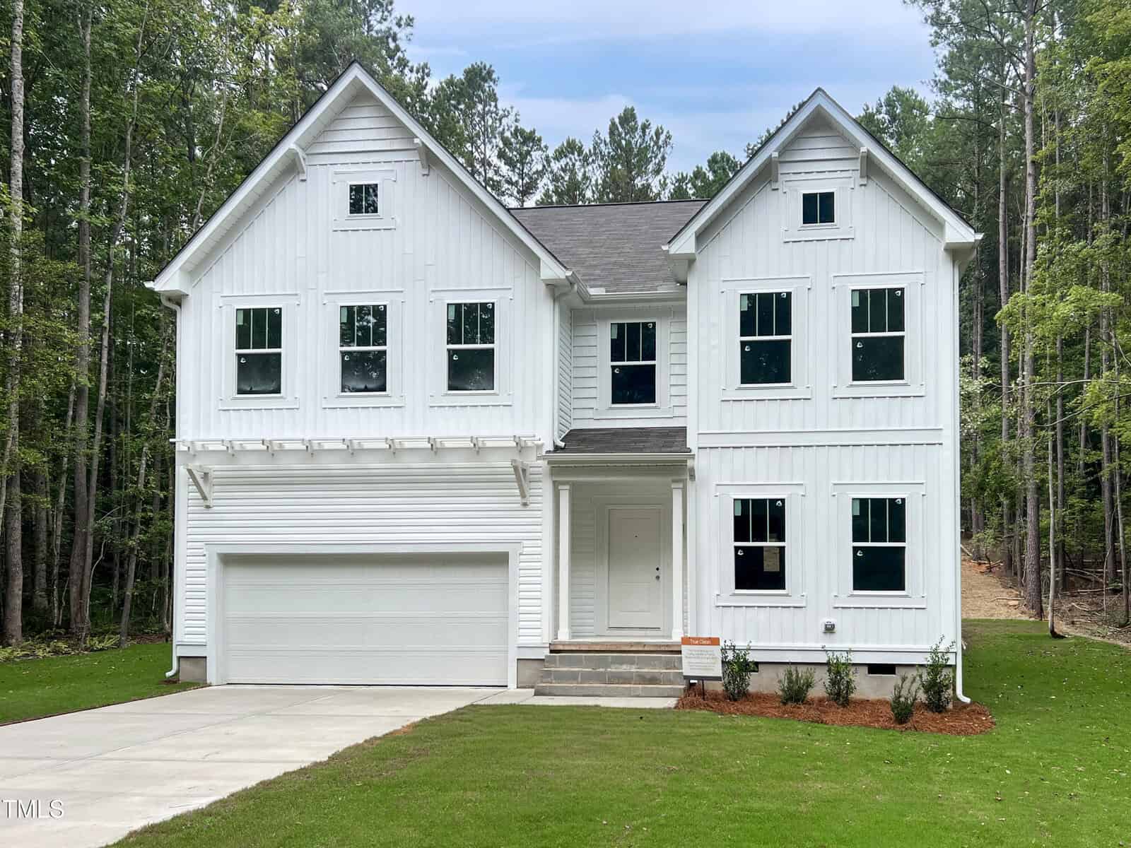 A modern two-story white house with a two-car garage, multiple windows, and a covered front porch, surrounded by green trees and a well-kept lawn.