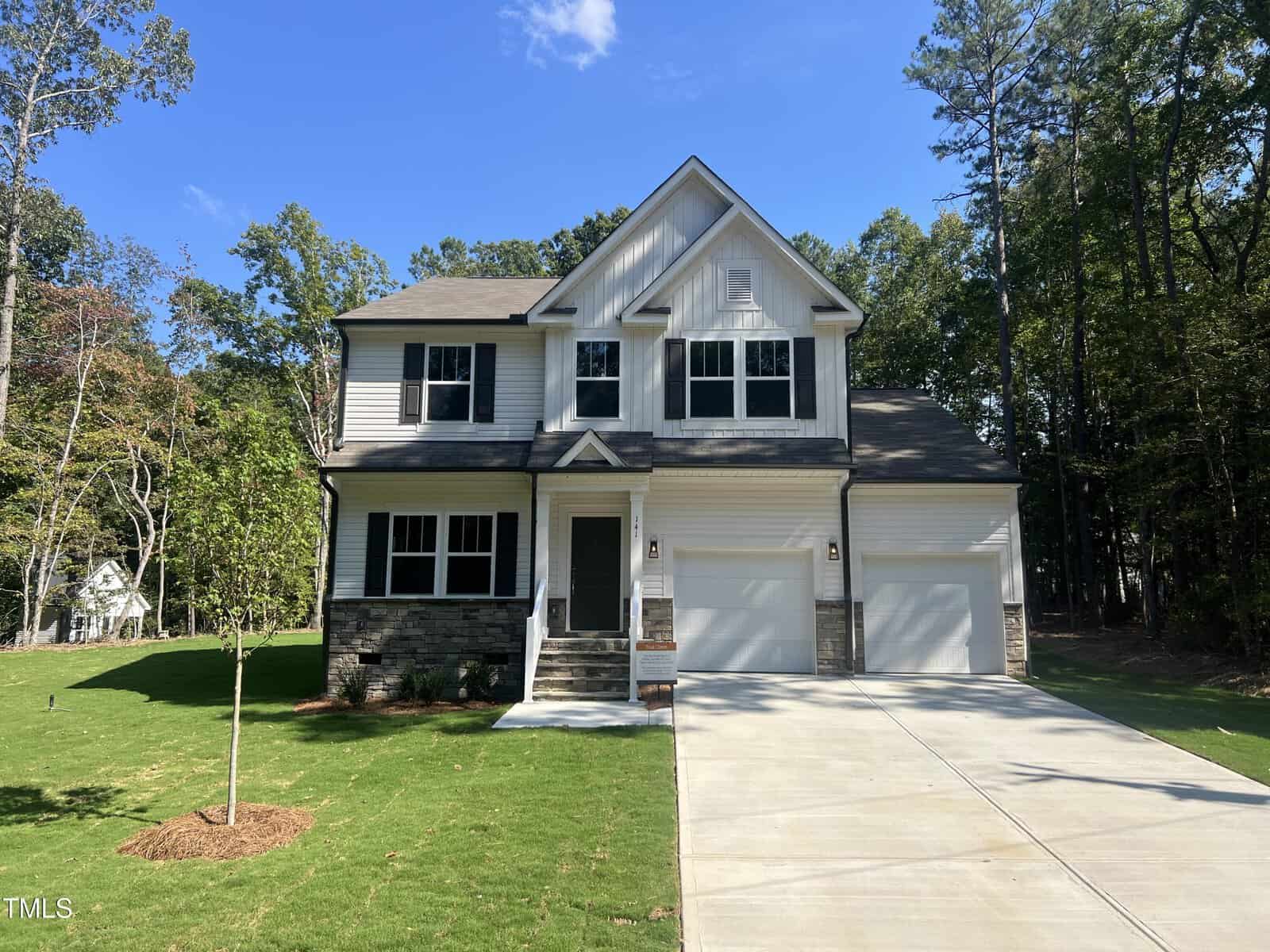 Nestled at 141 Shaman Drive 3012, Louisburg, this two-story suburban house with white siding, dark shutters, and a stone accent facade features a double garage, inviting front porch, and a well-kept lawn surrounded by tall trees under a clear sky.
