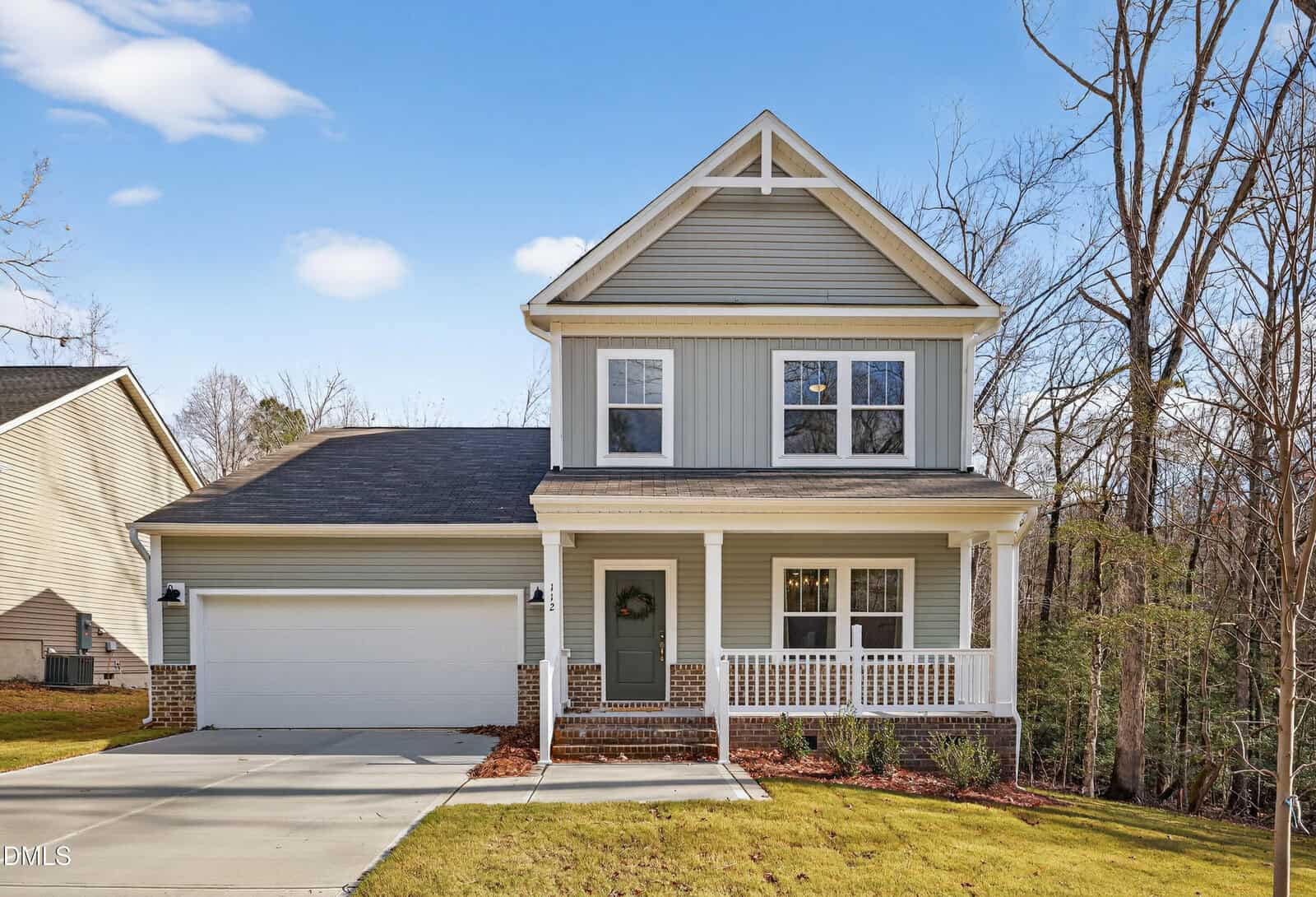 A two-story suburban house with gray siding, white trim, a covered front porch, and a two-car garage. The home sits on a well-kept lawn with trees and blue sky in the background.
