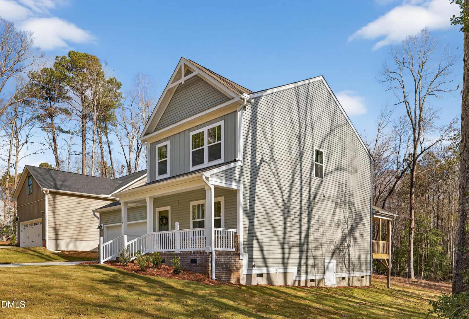 A two-story house with light gray siding, a white porch, and multiple windows, surrounded by trees and a grassy lawn under a blue sky with scattered clouds.