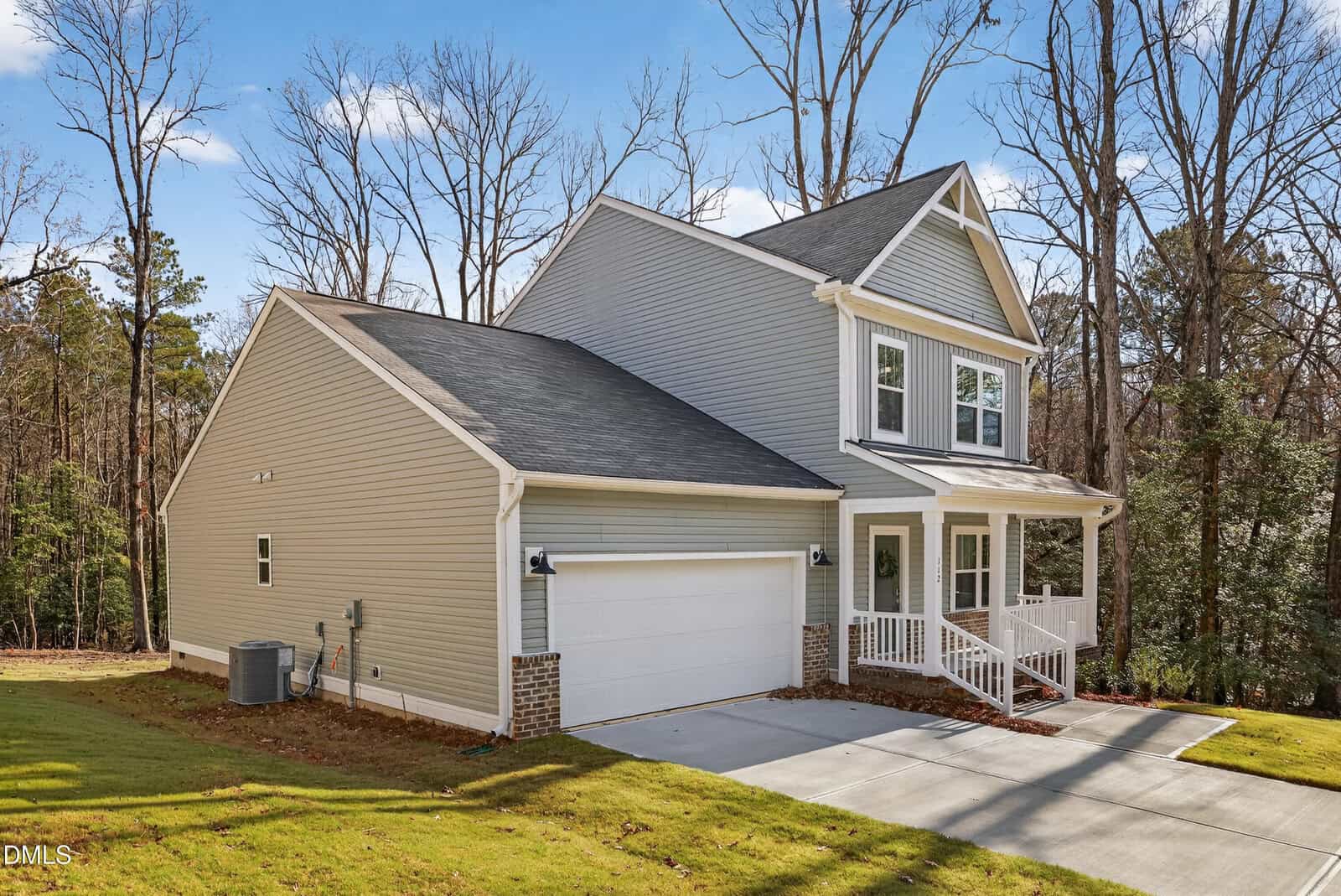 A modern two-story gray house with white trim, a double garage, and a front porch, set on a spacious lawn with trees and a forested background under a clear blue sky.