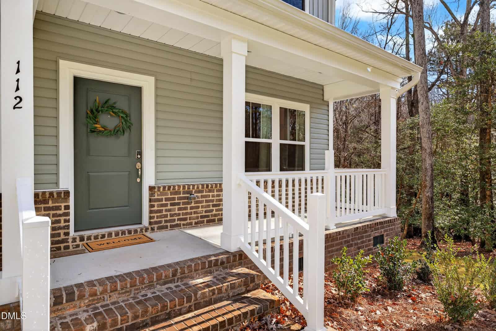 A green front door with a wreath leads to a brick porch with white railings, three steps, and a welcome mat. House number 112 is on the post. Shrubs and trees surround the entrance.