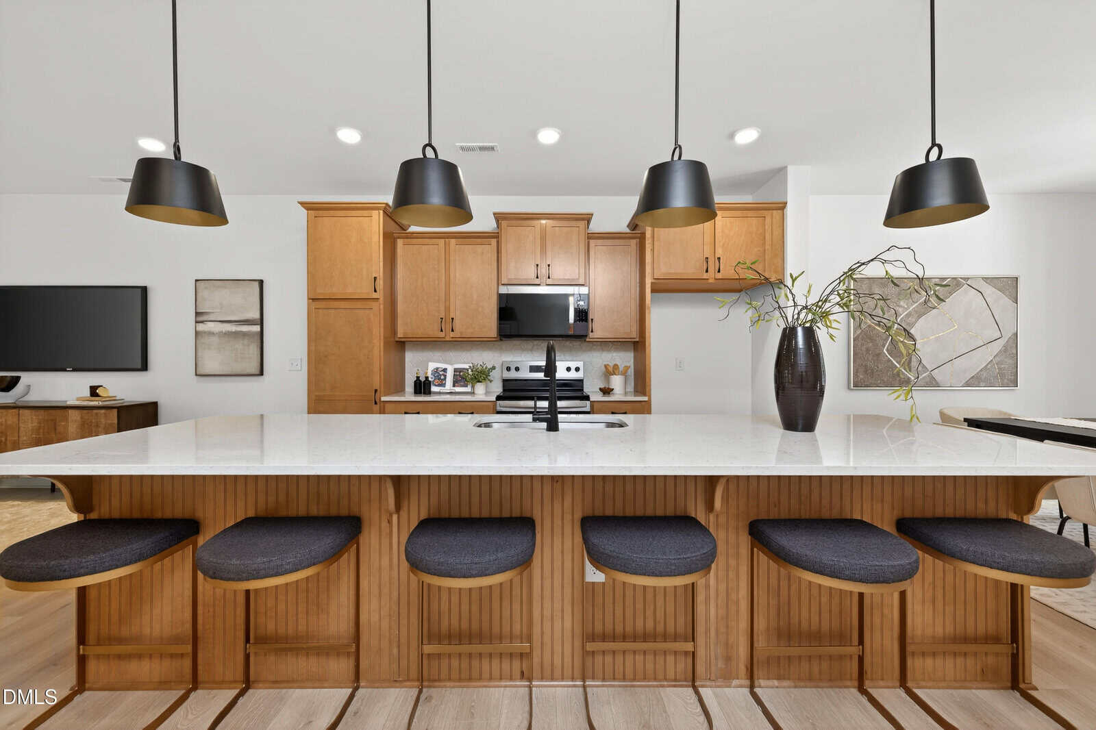 Modern kitchen with a large island, four cushioned stools, wood cabinetry, stainless steel appliances, three black pendant lights, and a tall vase with greenery on the countertop. Art and a TV are visible in the background.