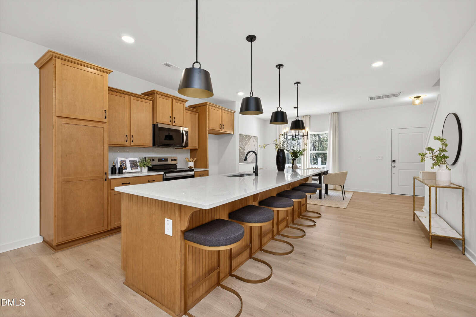 Modern kitchen with wooden cabinets, a large white marble island with four black barstools, stainless steel appliances, black pendant lights, and light wood flooring. Dining table and decor visible in the background.