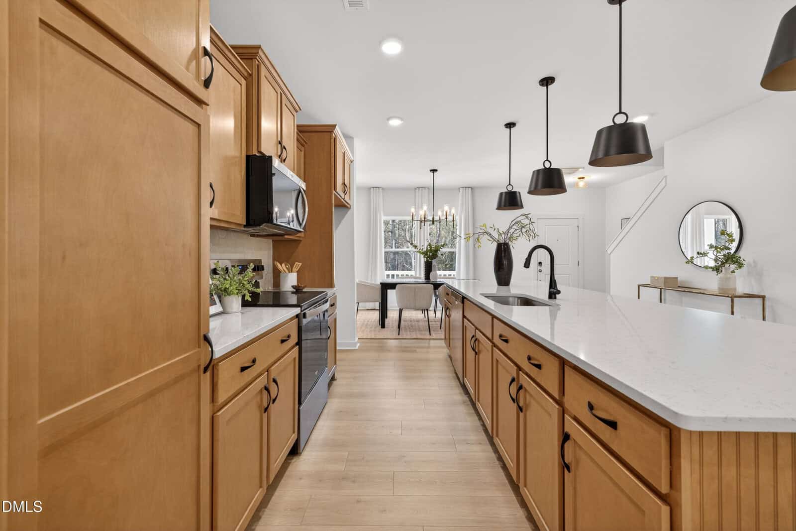 A modern kitchen with light wood cabinets, white countertops, a black oven, and a large island with a sink. Pendant lights hang above the island, and a dining area with a chandelier is visible in the background.