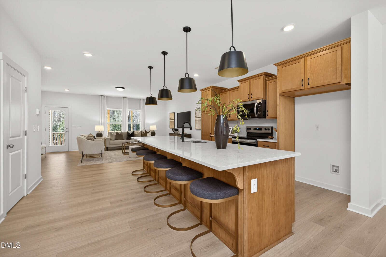 Modern open-concept kitchen with a long island, four barstools, wood cabinets, stainless steel appliances, pendant lights, and a living area with sofas and large windows in the background.