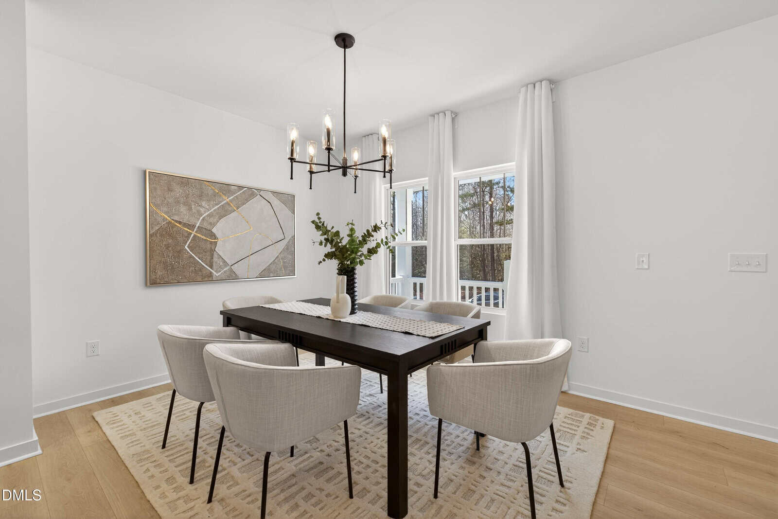 Bright dining room with a black rectangular table, six white cushioned chairs, a modern chandelier, large abstract wall art, and a window with white curtains letting in natural light. A vase with greenery sits on the table.