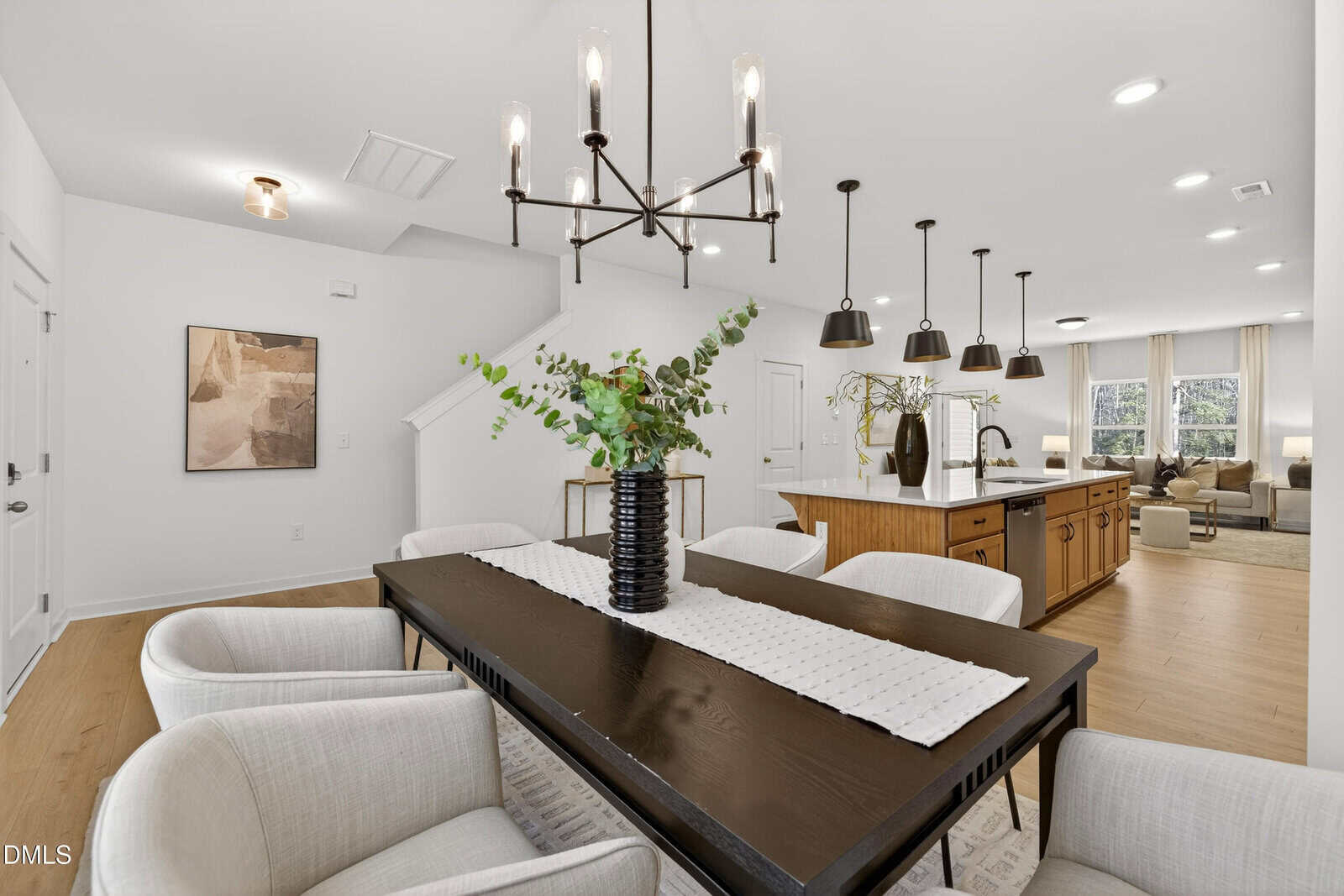 Modern open-concept dining and kitchen area with a black rectangular table, beige upholstered chairs, a black vase with greenery, pendant lights over a kitchen island, and neutral decor throughout.