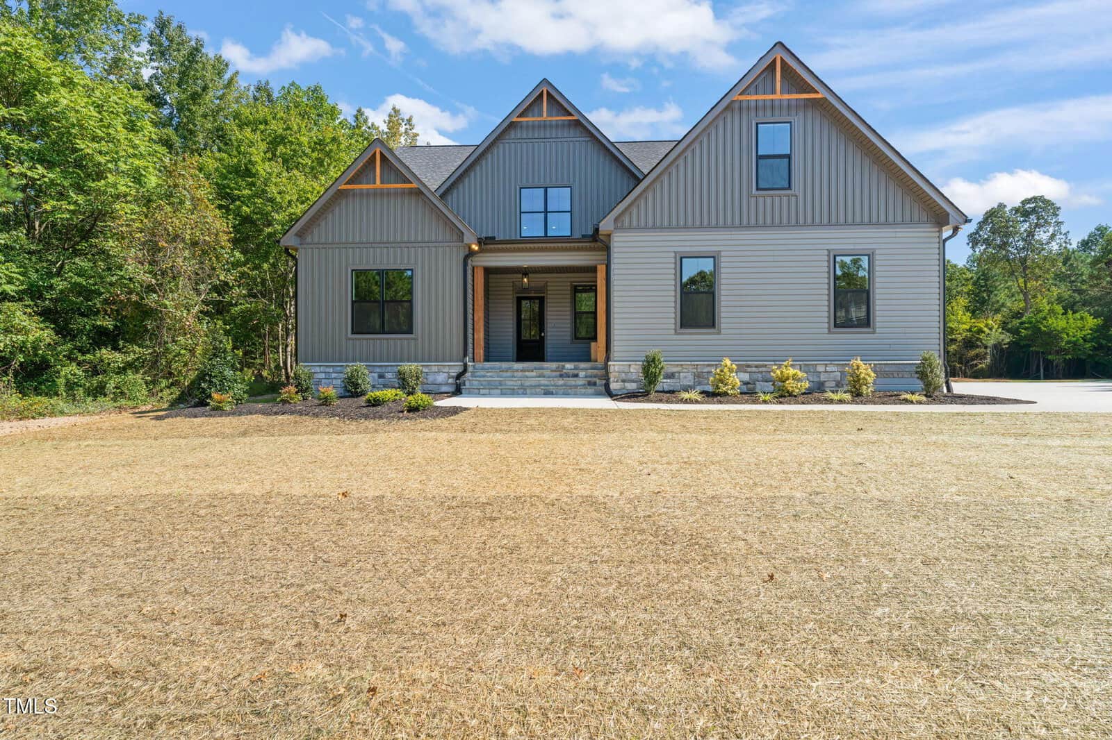 Modern two-story house with gray vertical siding, large windows, a covered front porch, and gable roof accents located at 360 Pilot Ridge Road in Zebulon. The front yard features new landscaping, a stone walkway, and patchy grass, all surrounded by trees and set against a bright blue sky.