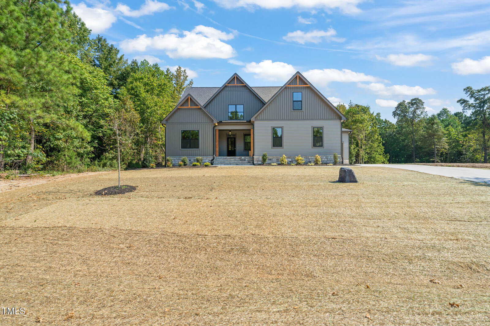 A modern, light gray two-story house with gabled roofs sits on a large, mostly bare lawn bordered by trees. A new sapling and a cut tree stump are visible in the front yard under a blue sky with clouds.