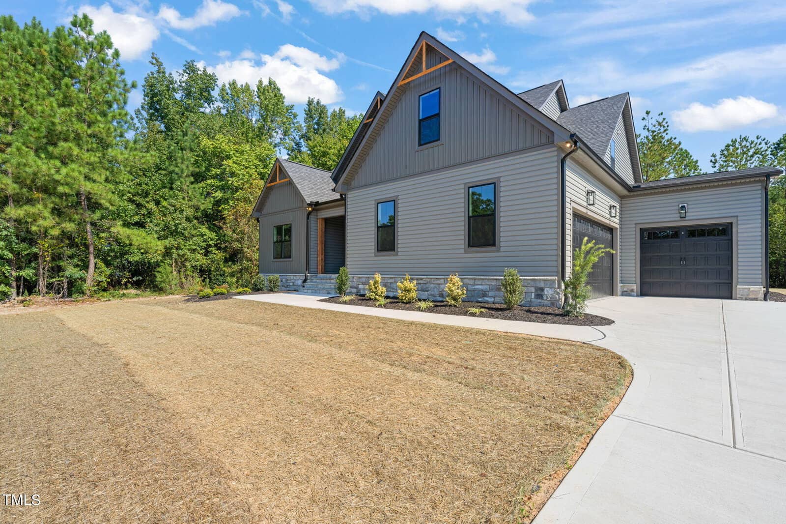 Modern gray house with gable roof, stone accents, attached garage, and a new driveway. The front yard has brown patchy grass and small shrubs. Trees and greenery are visible in the background under a blue sky with clouds.