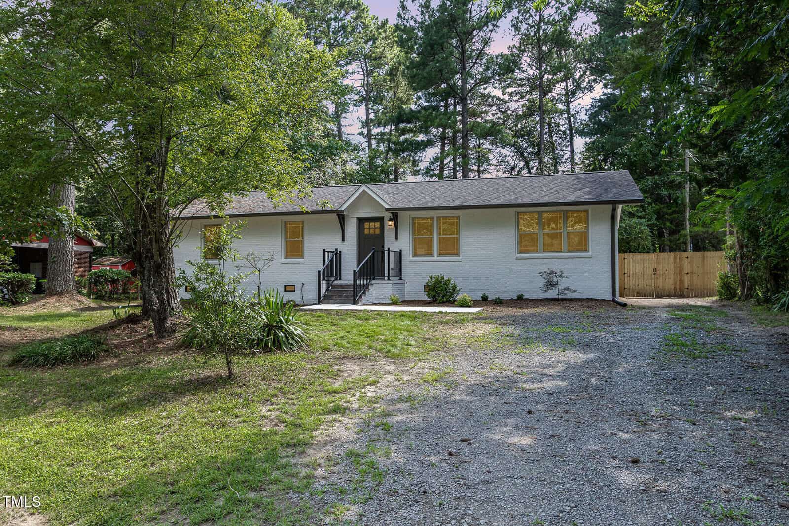 A single-story white house at 605 26th Street, Butner, with yellow-trimmed windows, a black front door, metal railings, and a gravel driveway, surrounded by trees and greenery.