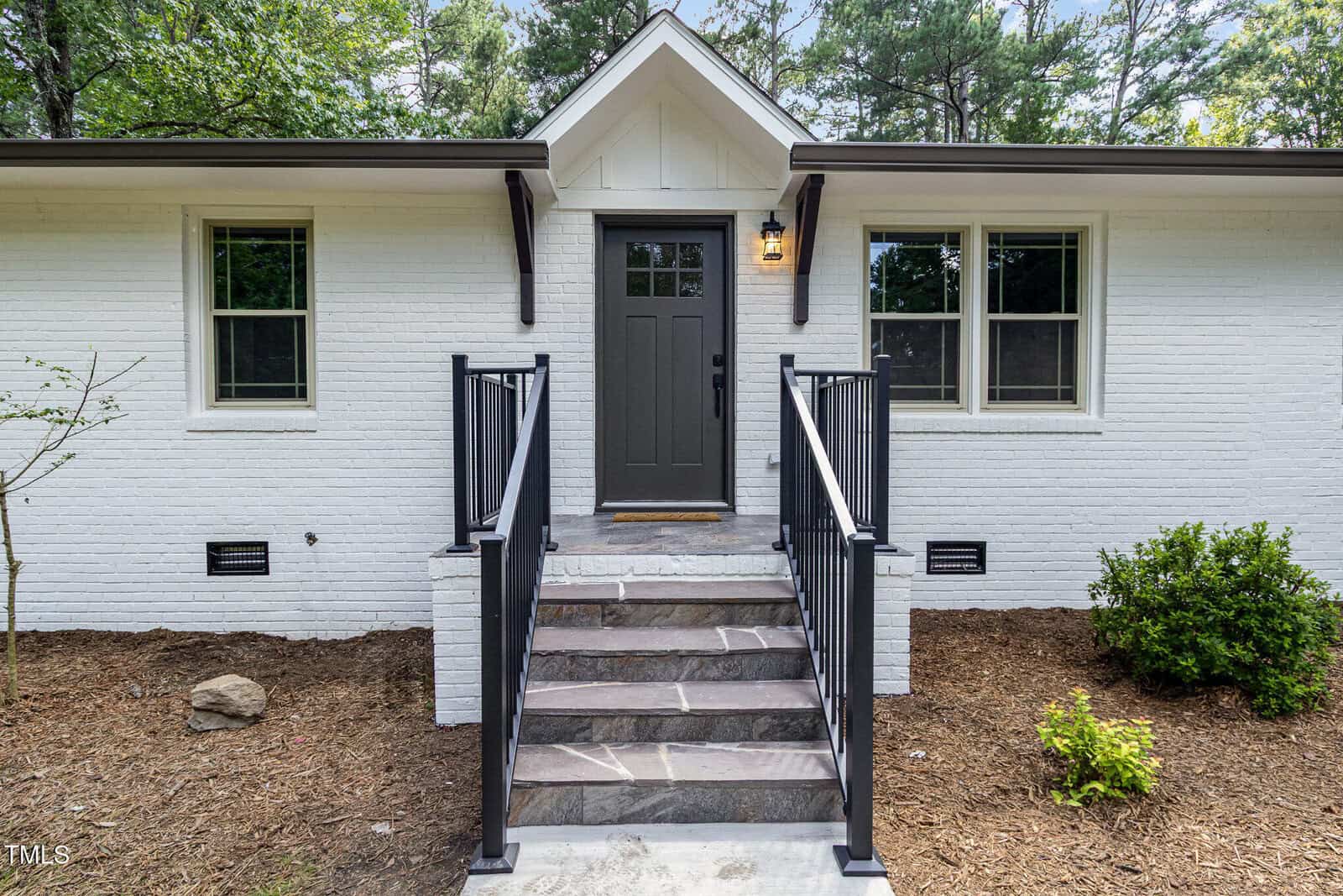 A small white brick house with a dark gray front door, two windows on either side, black railings along stone steps leading to the entrance, and simple landscaping with mulch and shrubs.
