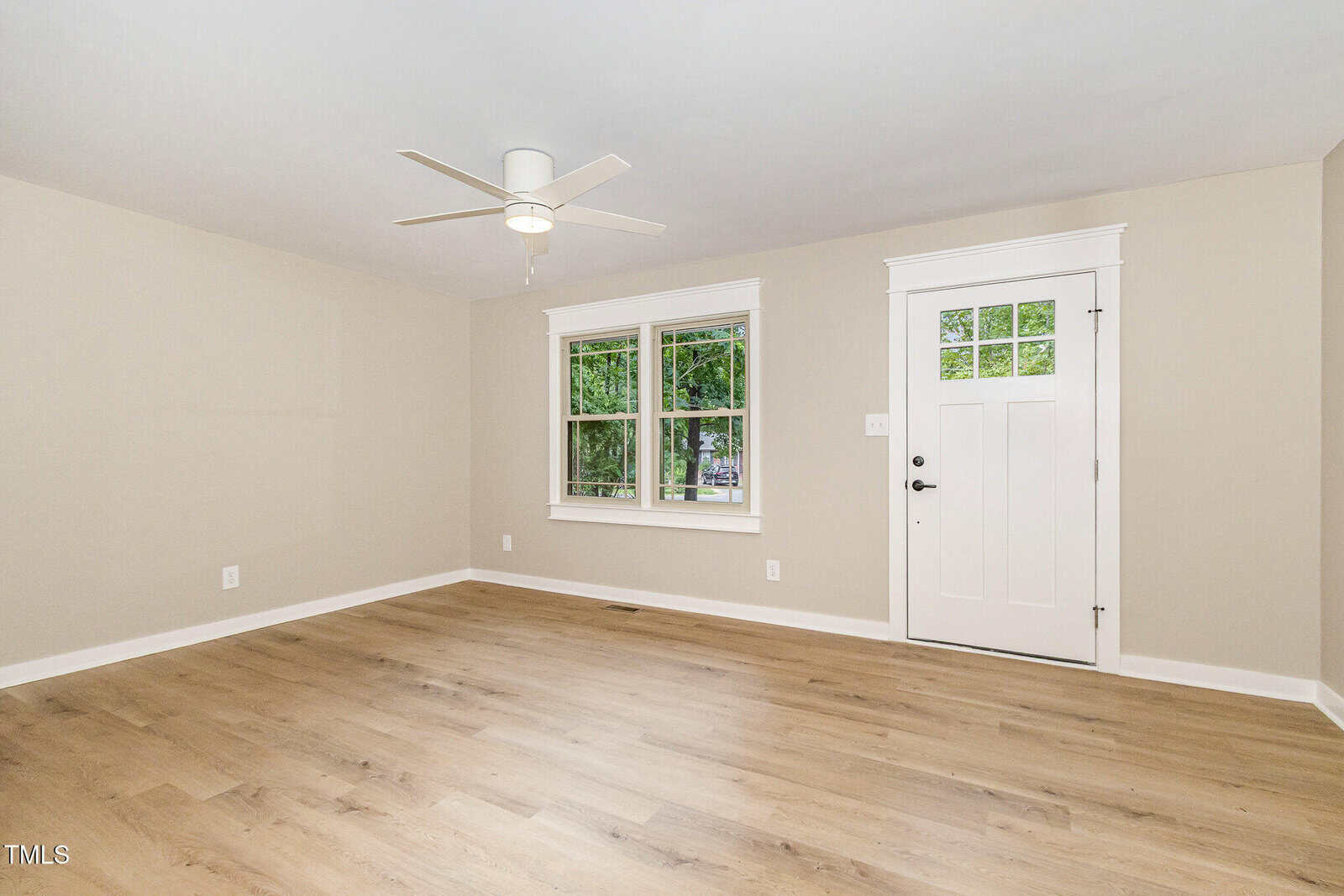 A bright, empty room with light wood flooring, beige walls, a white ceiling fan, a white door, and two windows showing green trees outside. White trim frames the windows, door, and baseboards.