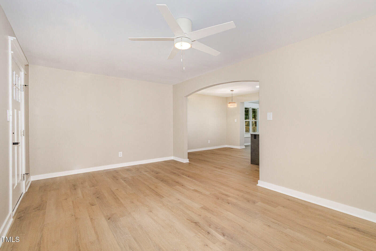 A spacious, empty room with light wood flooring, beige walls, a white ceiling fan, and an arched doorway leading to another room. Recessed lighting and baseboards are visible.