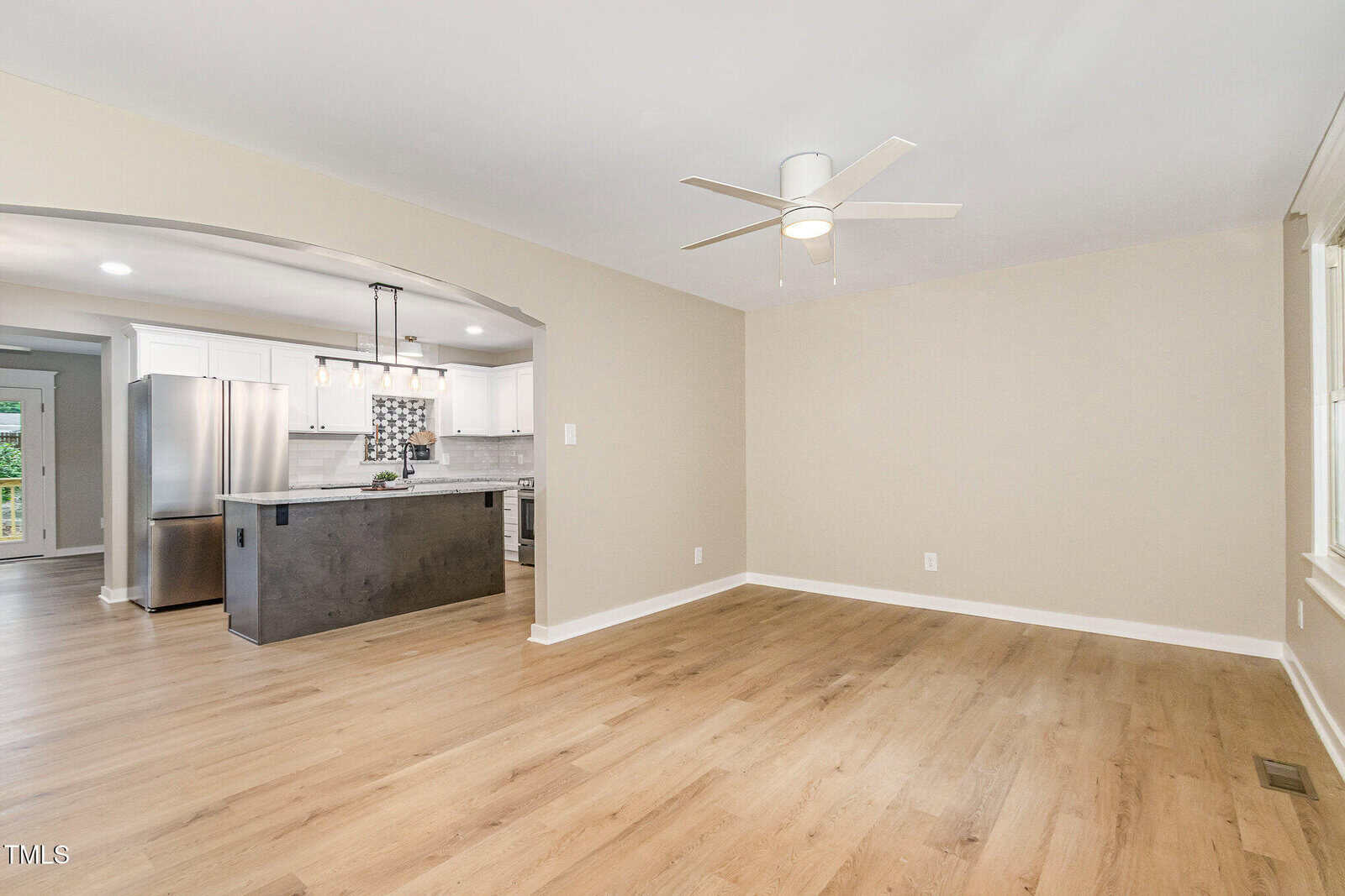 A spacious, empty room with light wood floors, beige walls, a white ceiling fan, and an open view of a modern kitchen with white cabinets, black island, hanging lights, and stainless steel appliances.