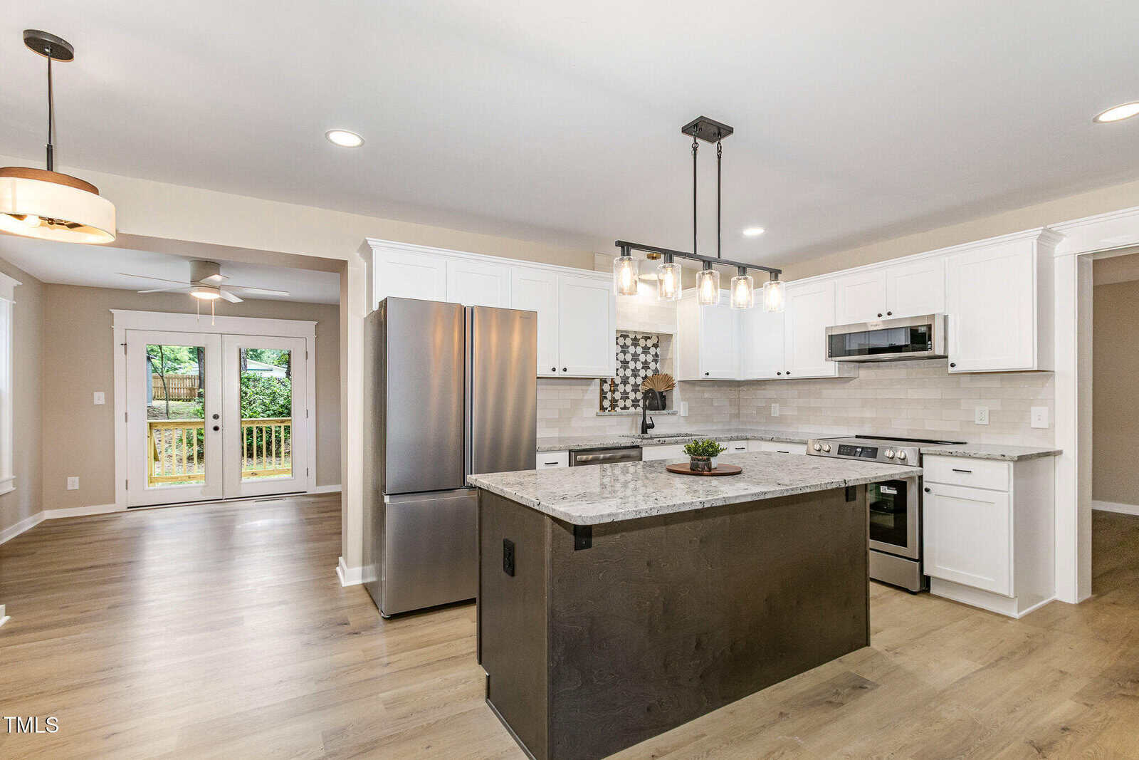 A modern kitchen with white cabinets, stainless steel appliances, a central island with granite countertop, pendant lighting, light wood flooring, and a view of a backyard through glass double doors.