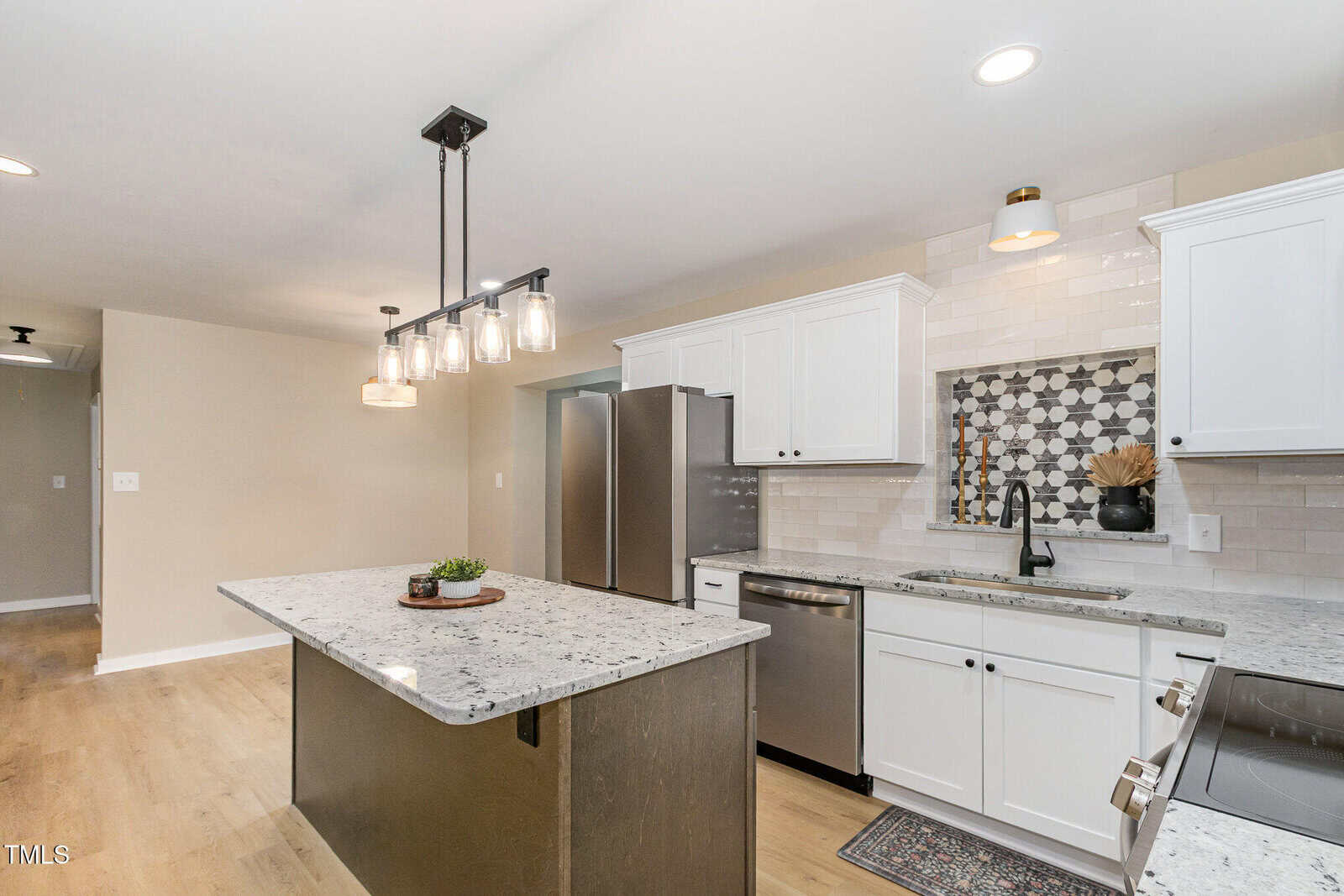 A modern kitchen features white cabinets, stainless steel appliances, a light granite island with pendant lights above, wood flooring, and a decorative tile backsplash behind the sink.
