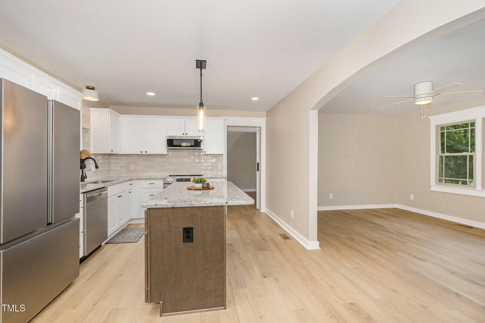 A modern kitchen with white cabinets, stainless steel appliances, an island with a dark base, pendant lighting, and light wood flooring, adjacent to an open living area with large windows.