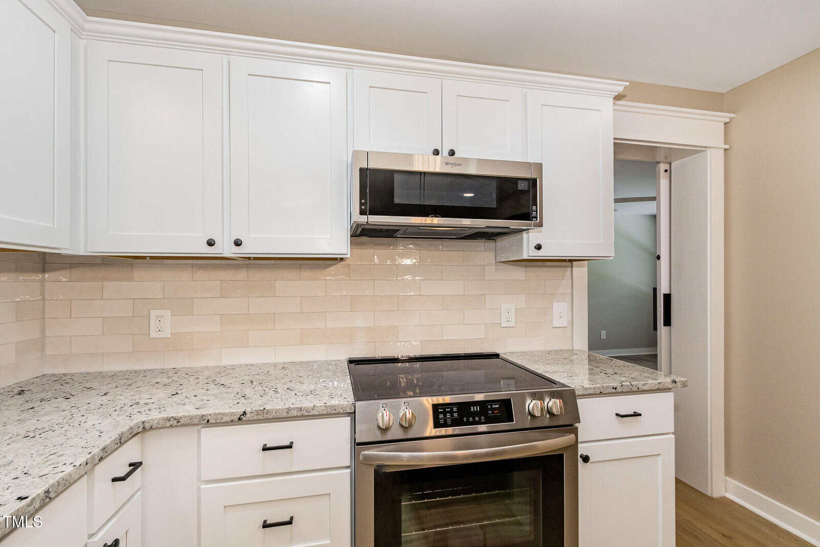 Modern kitchen with white cabinets, a stainless steel oven and microwave, granite countertops, beige subway tile backsplash, and light wood floors. A doorway leads to another room in the background.