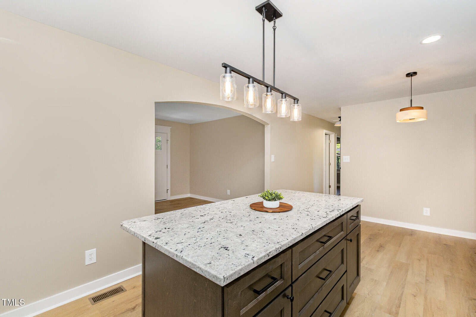 Modern kitchen with a large island featuring a white granite countertop, dark cabinetry, pendant lighting above, light wood floors, and beige walls with an arched doorway.