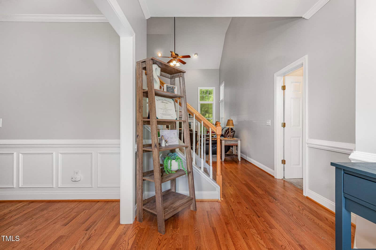 A wooden bookshelf stands near a staircase with wood steps and white railing in a home with light gray walls, white trim, and hardwood floors. A door leads to another room on the right.