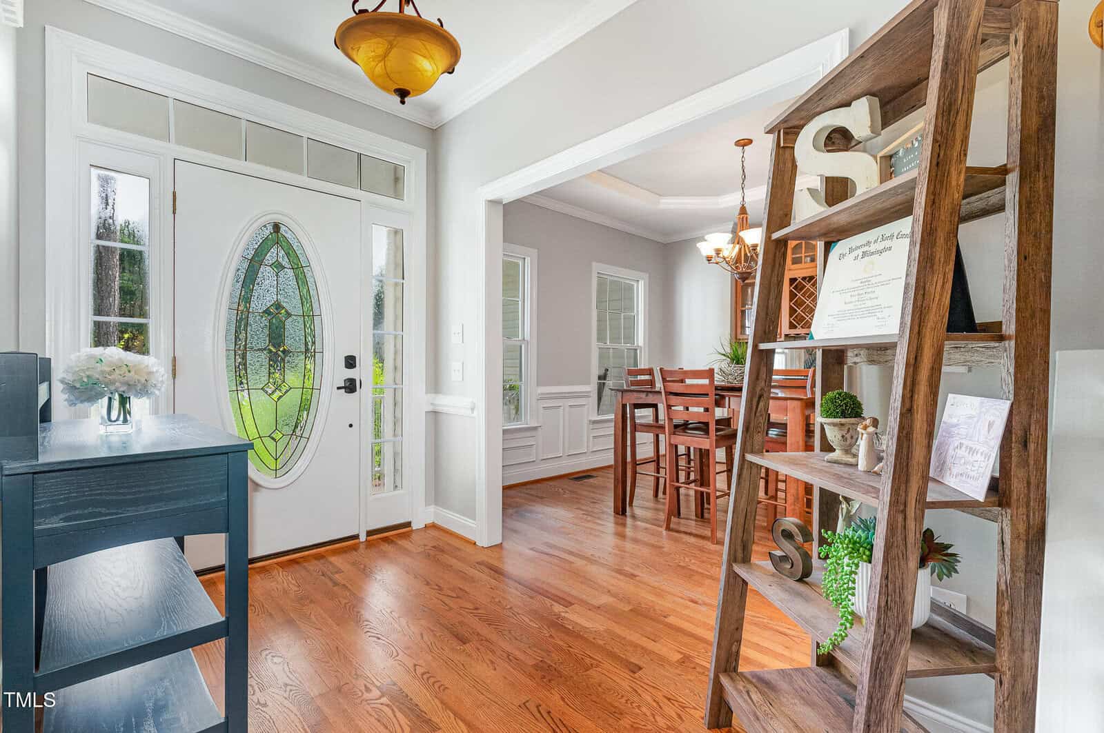 A bright entryway with a glass-paneled front door, wooden floors, a blue console table, and a tall wooden shelf. A dining room with a wooden table and chairs is visible through a large doorway.