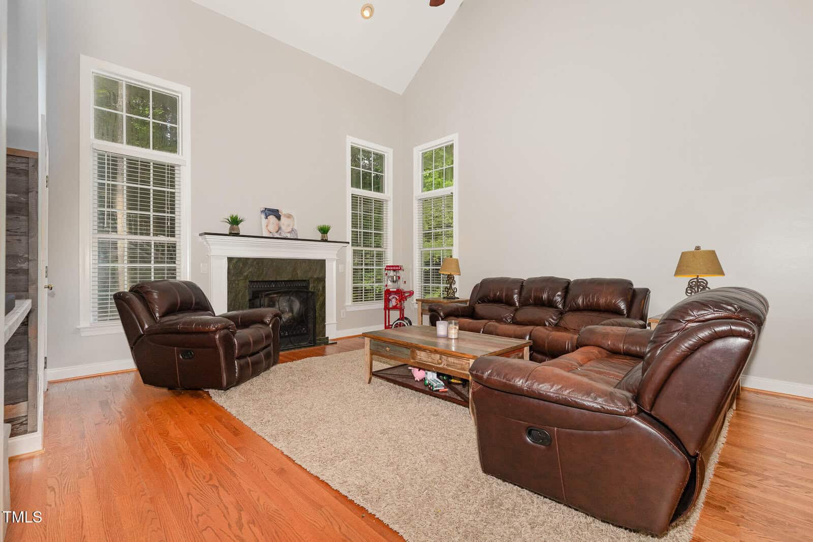 A living room with brown leather sofas, a matching recliner, a wooden coffee table on a beige rug, hardwood floors, a fireplace with a mantle, and three tall windows letting in natural light.